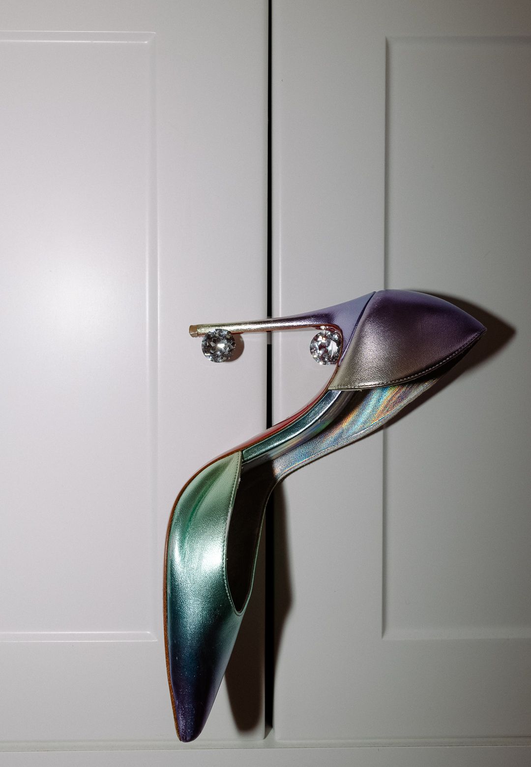 Iridescent multicolor bridal heels with crystal embellishments displayed against a white paneled door.