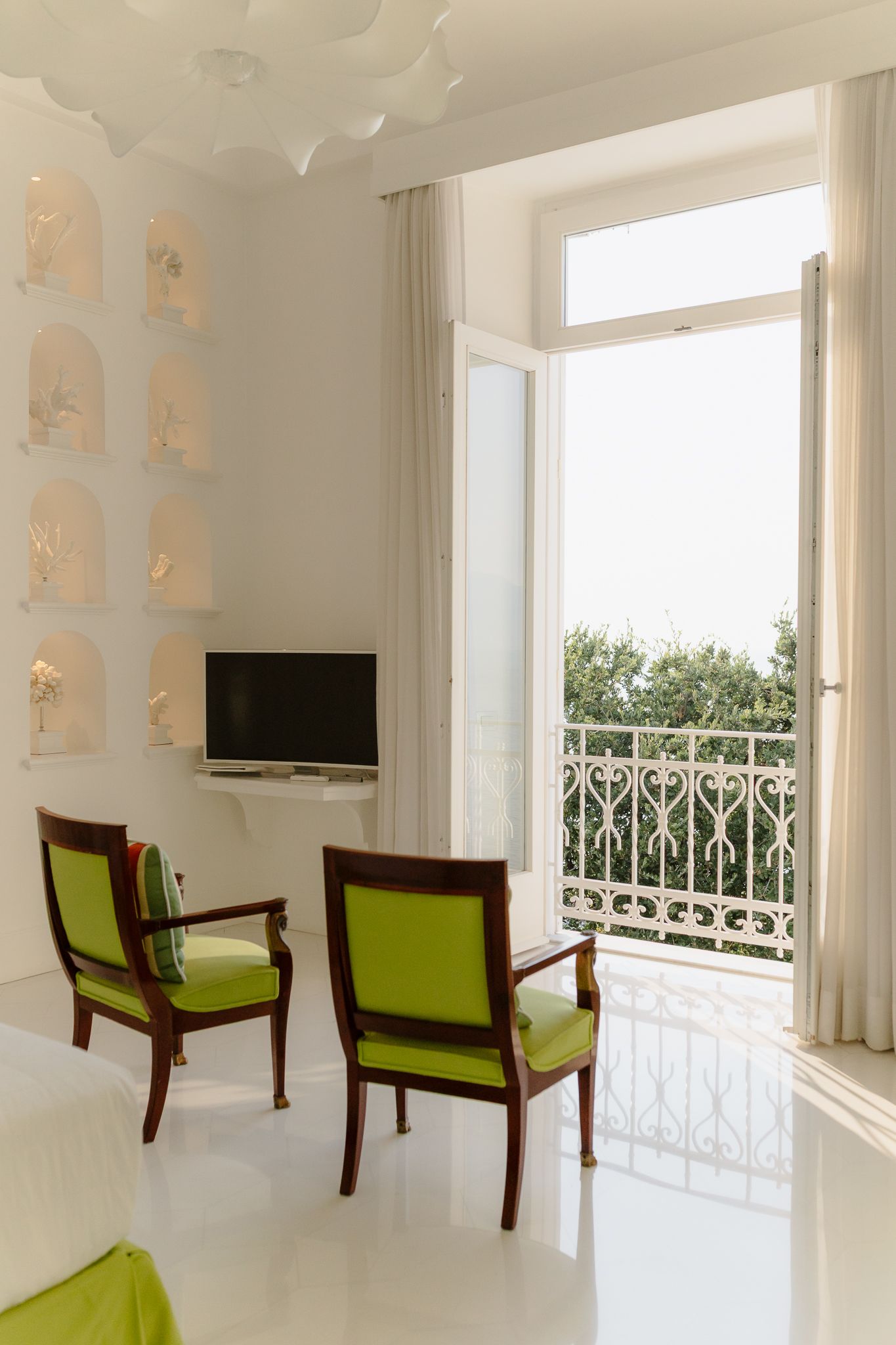 Bright white room with green chairs, wall niches, TV, and open balcony with garden view.