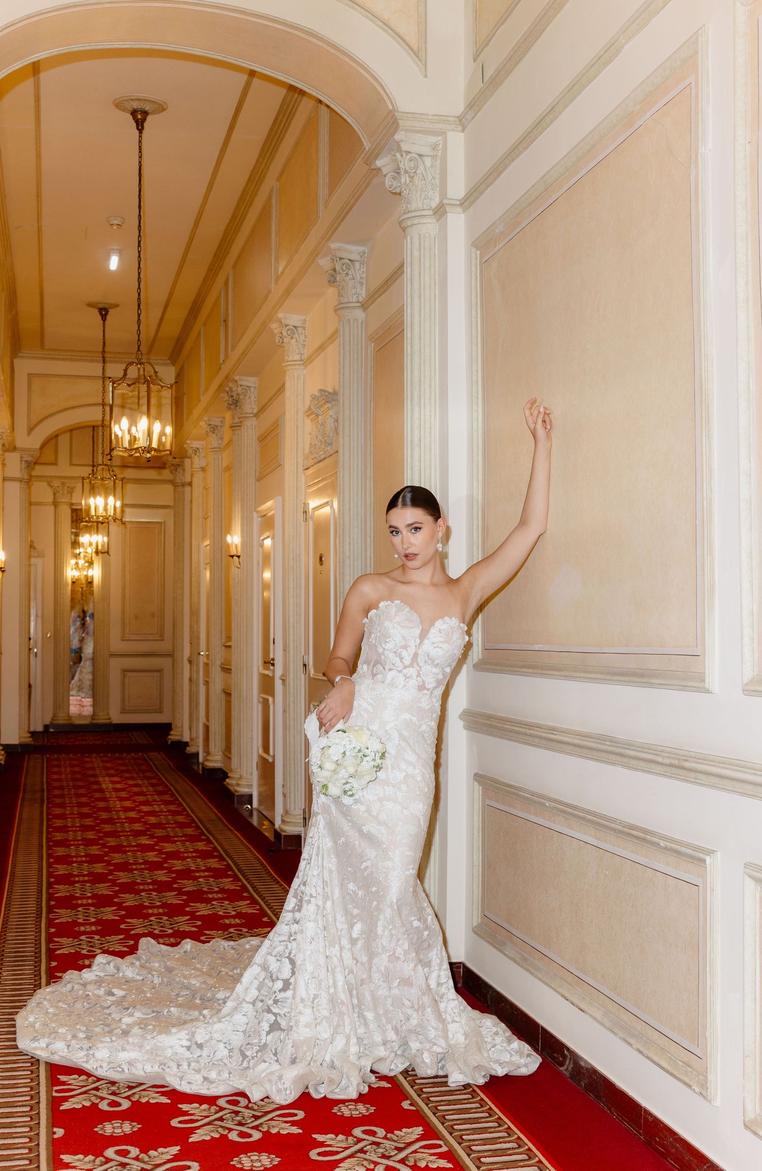 Stunning bride in strapless lace mermaid wedding gown posing in grand hotel corridor with red carpet.