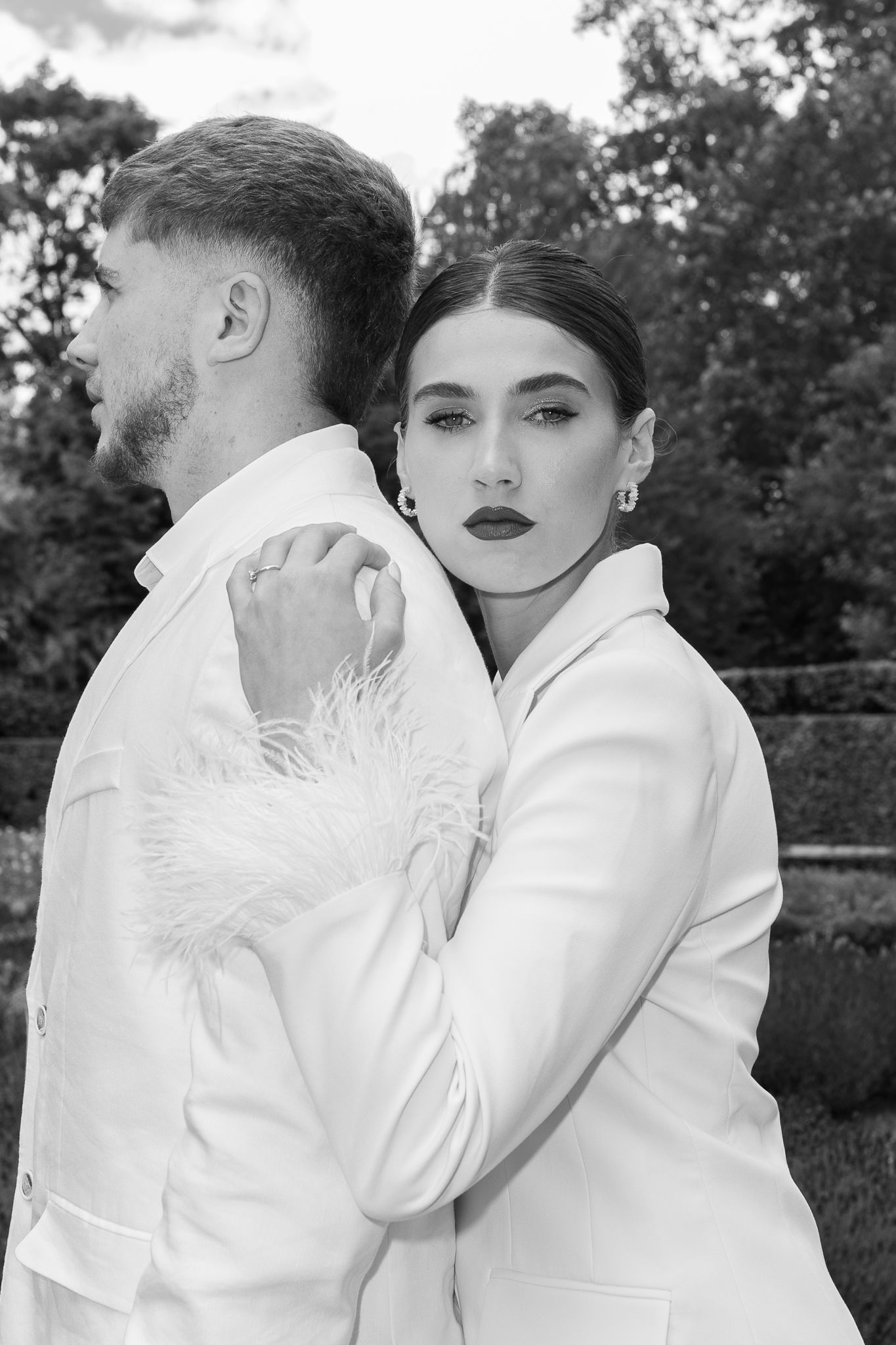 Black and white photo of an elegant couple in white suits embracing outdoors, woman gazing at camera with bold makeup.