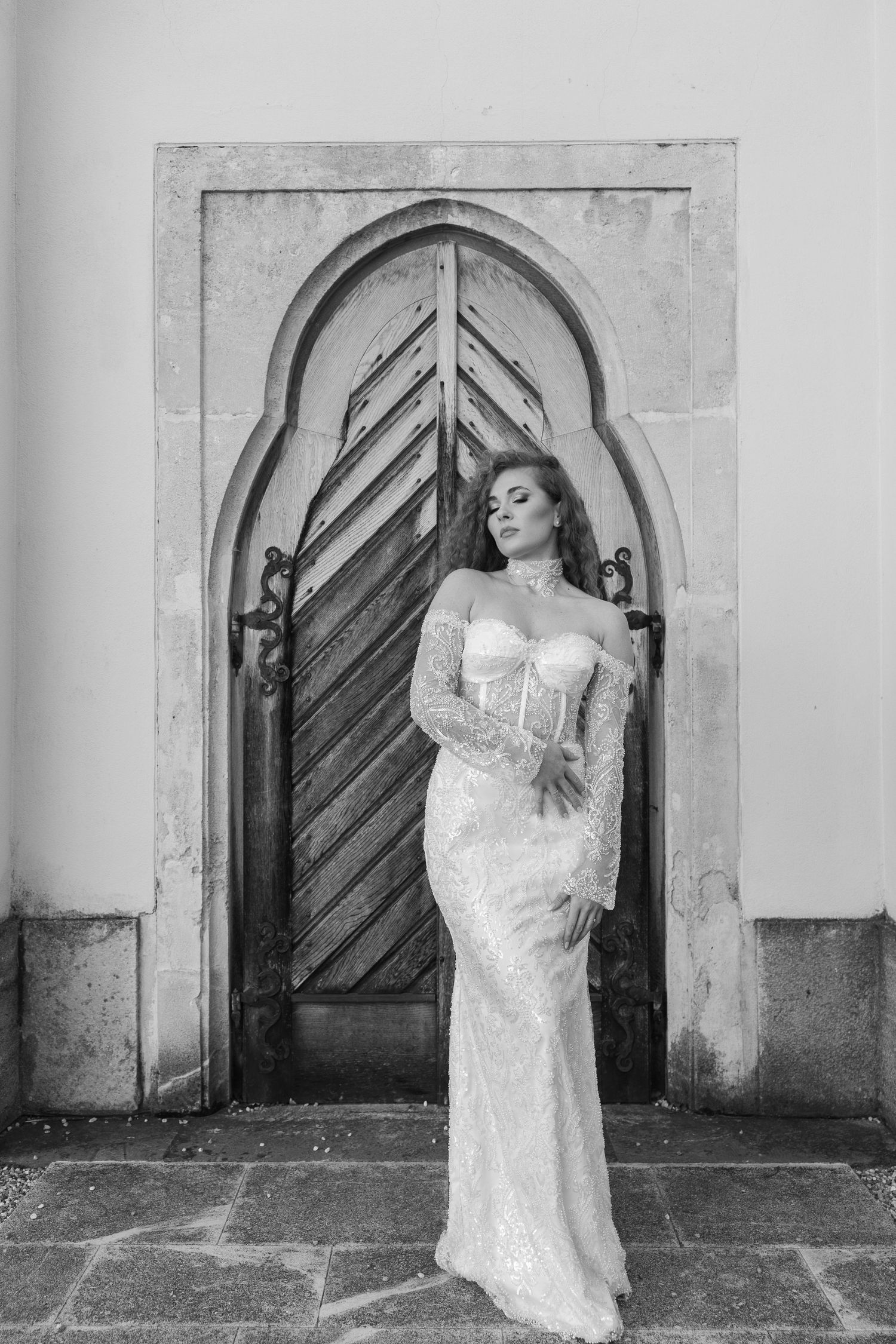 Bride in off-shoulder sequin wedding gown posing before an ornate arched wooden door.