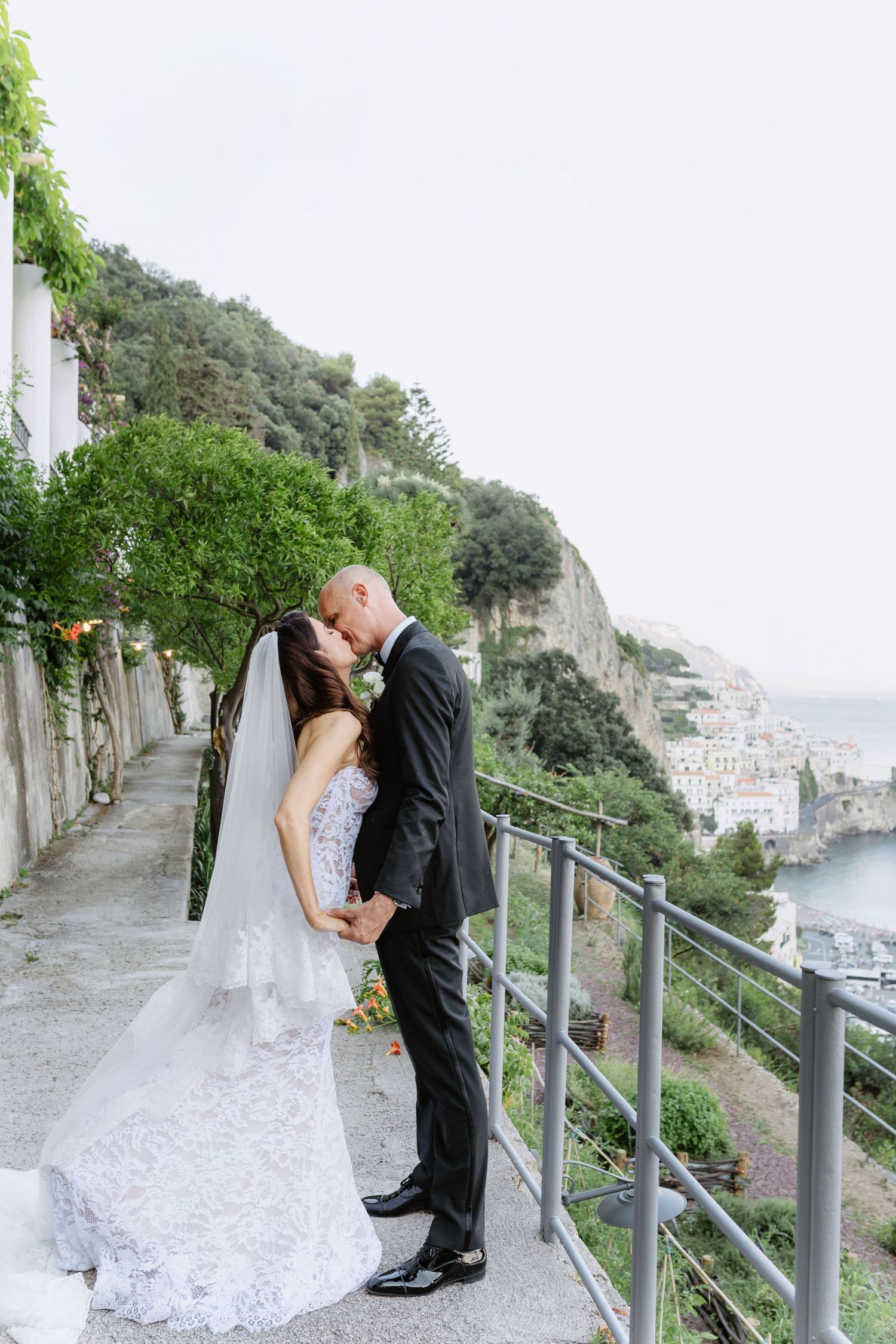 Bride and groom share a romantic kiss on a scenic Amalfi Coast terrace, with cliffside town views behind them.