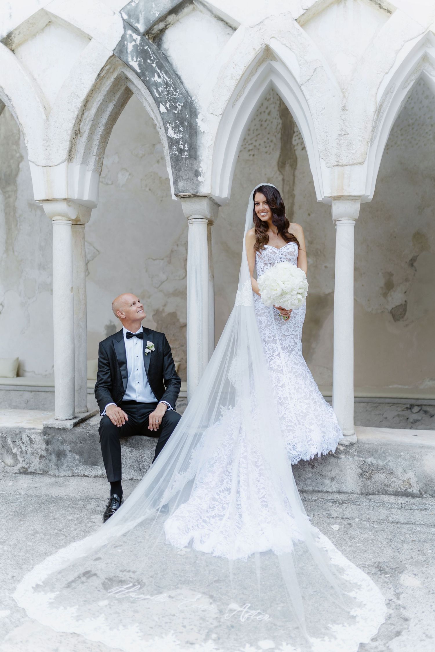 Bride in lace mermaid gown with long veil stands beside groom in tuxedo among Gothic arches at elegant wedding venue.