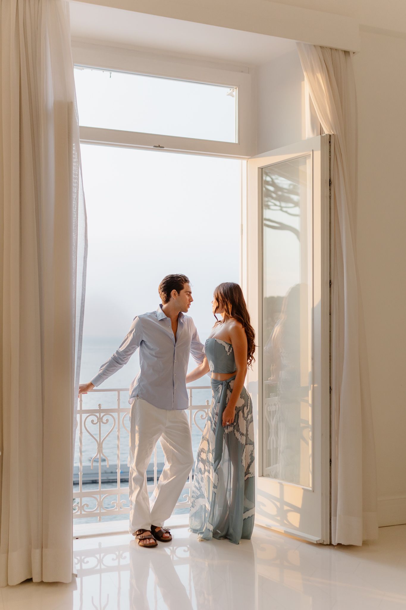 Couple stands in sunlit open doorway with ocean view balcony in romantic coastal villa.