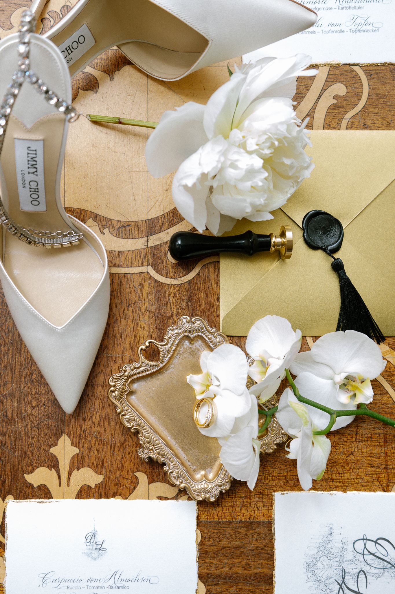 Jimmy Choo bridal shoes with white orchids, gold envelope, wax seal, and wedding stationery flatlay.