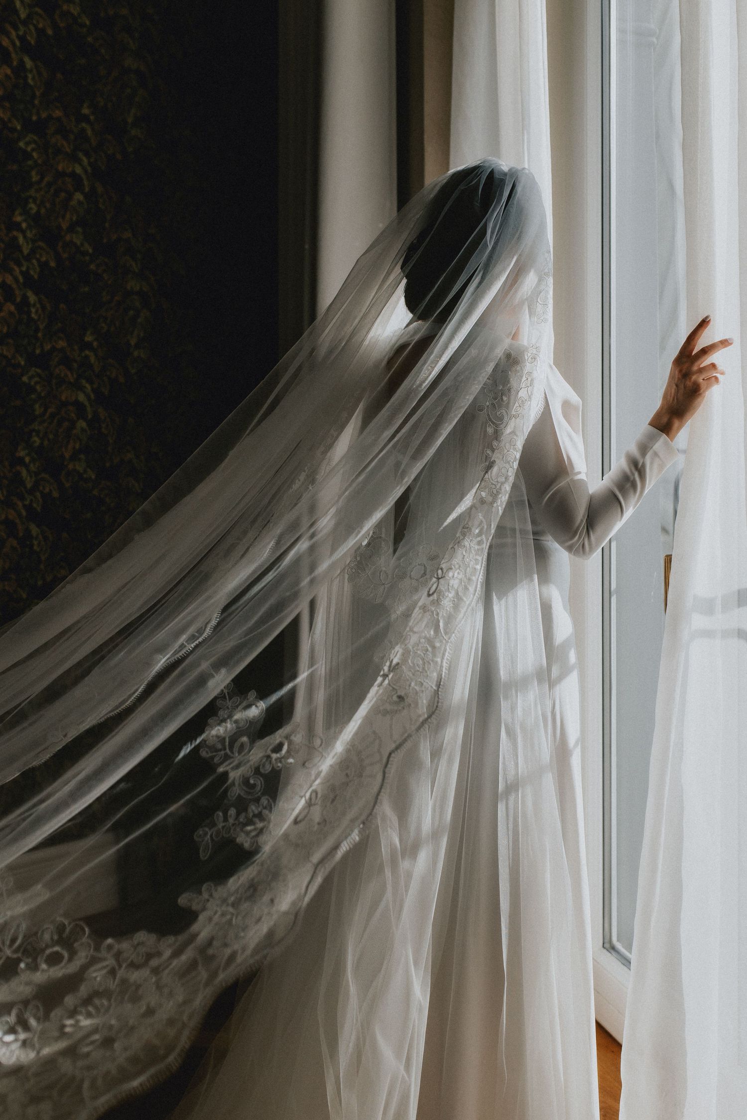 Bride in lace wedding dress with long cathedral veil gazing out a bright window.