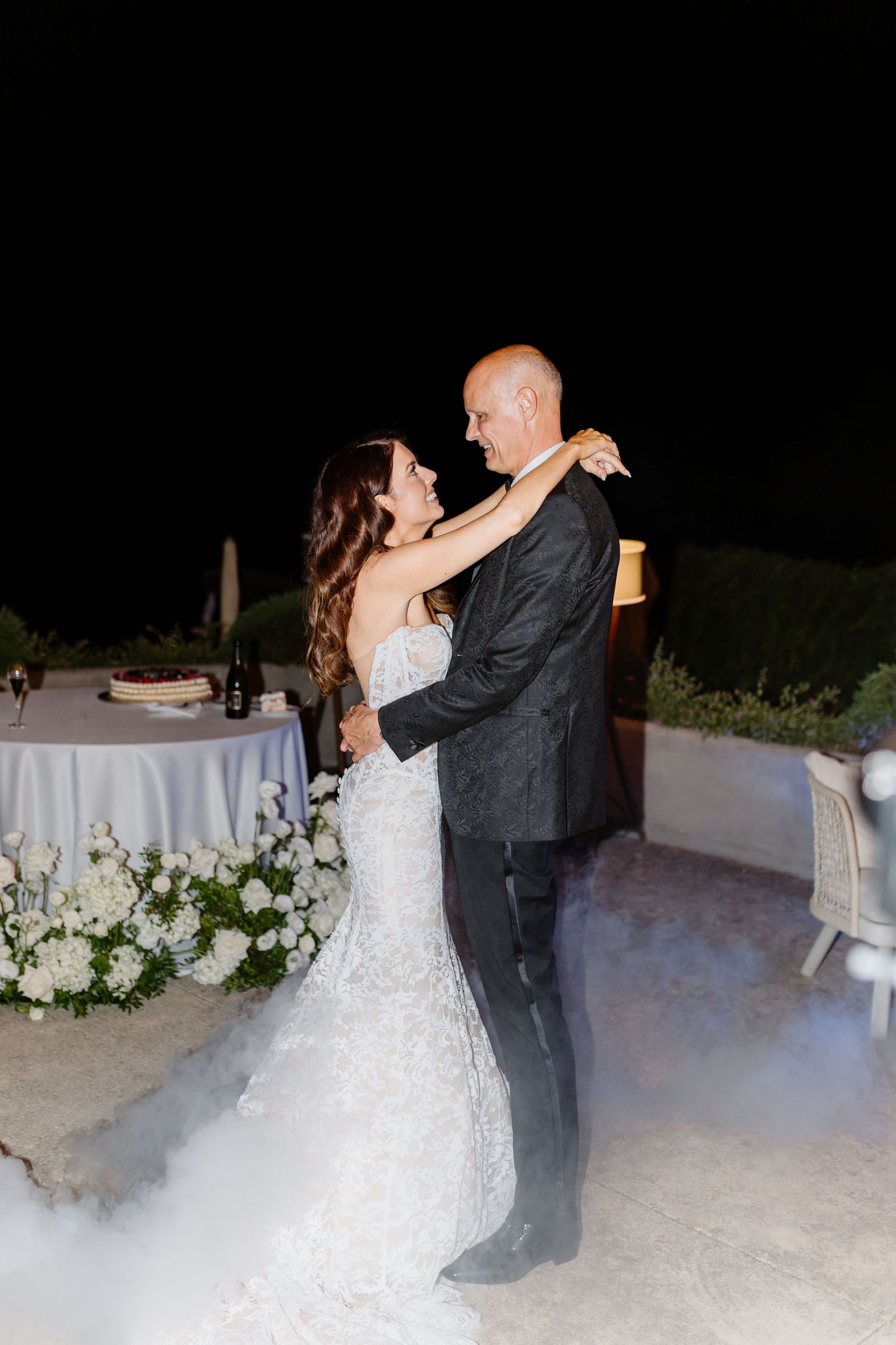 Bride and groom share their first dance outdoors at night, surrounded by white floral arrangements and a dreamy fog effect.