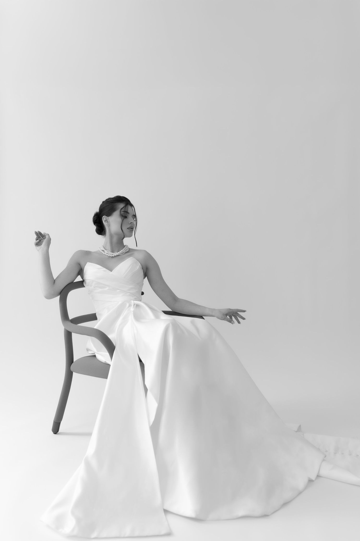 Black and white portrait of bride in strapless satin wedding gown seated gracefully on a wooden chair.