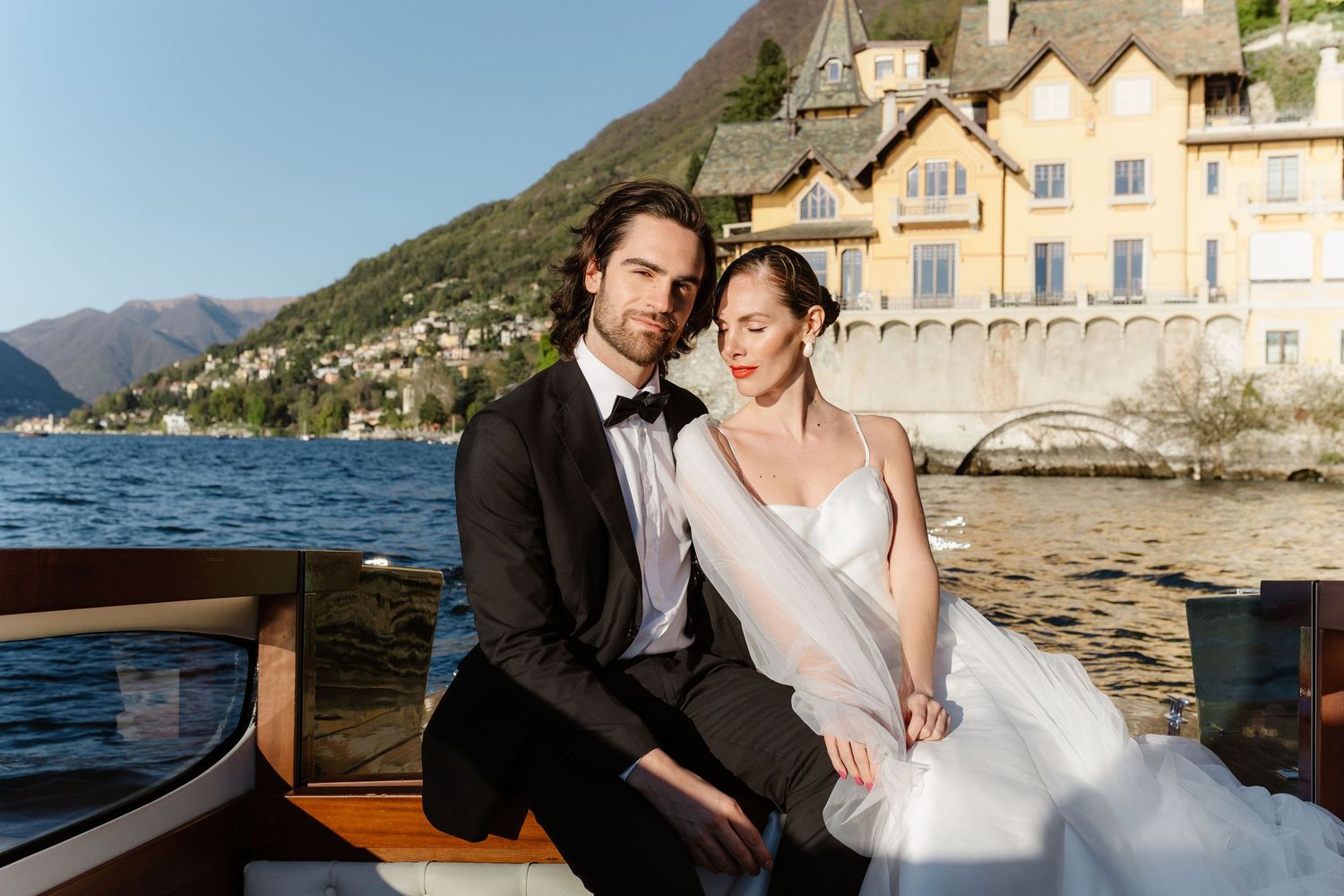 Bride and groom in wedding attire sitting together on a classic wooden boat by a lakeside villa.