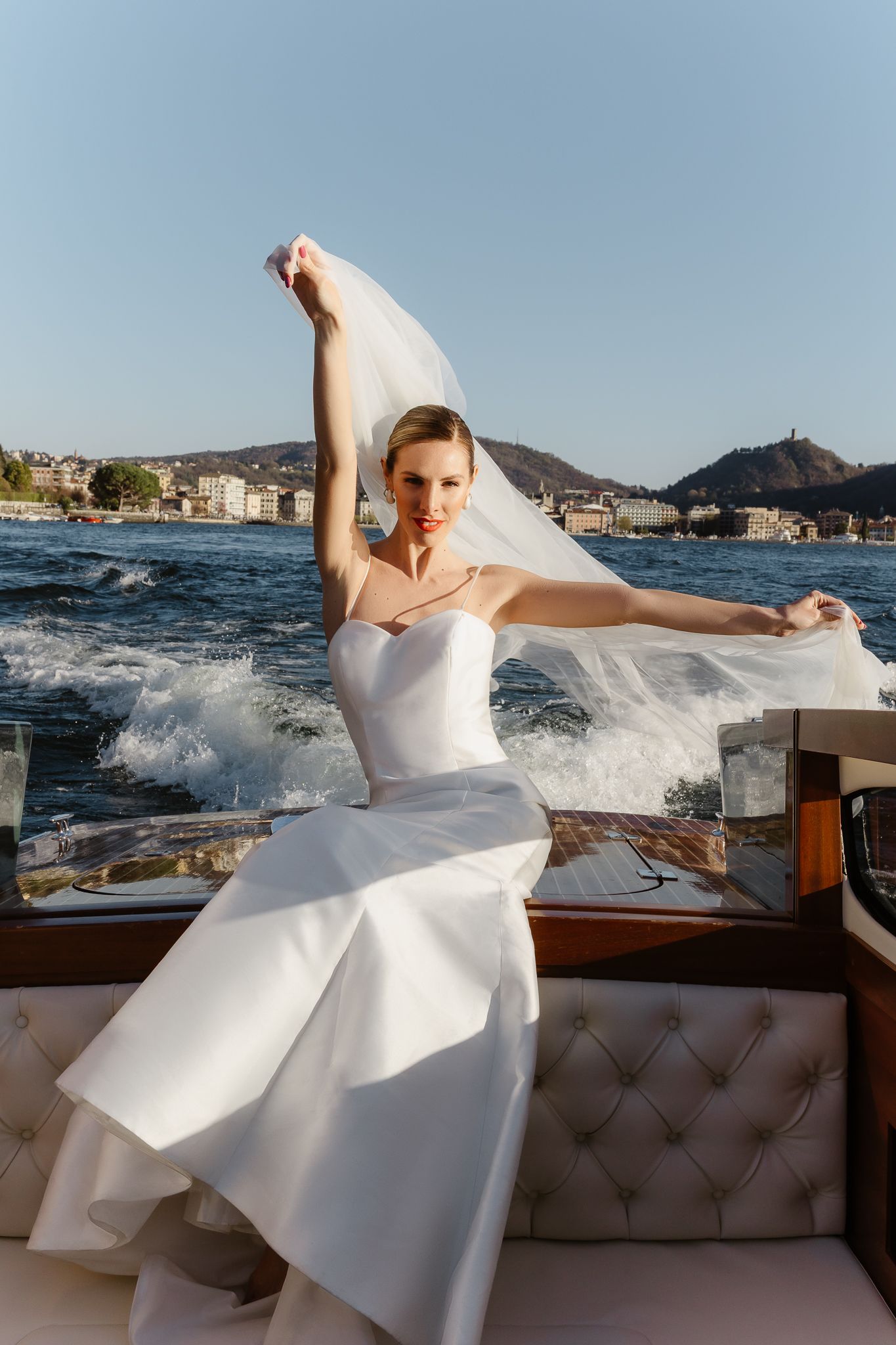 Bride in white gown holds veil on luxury boat with mountain lake backdrop.