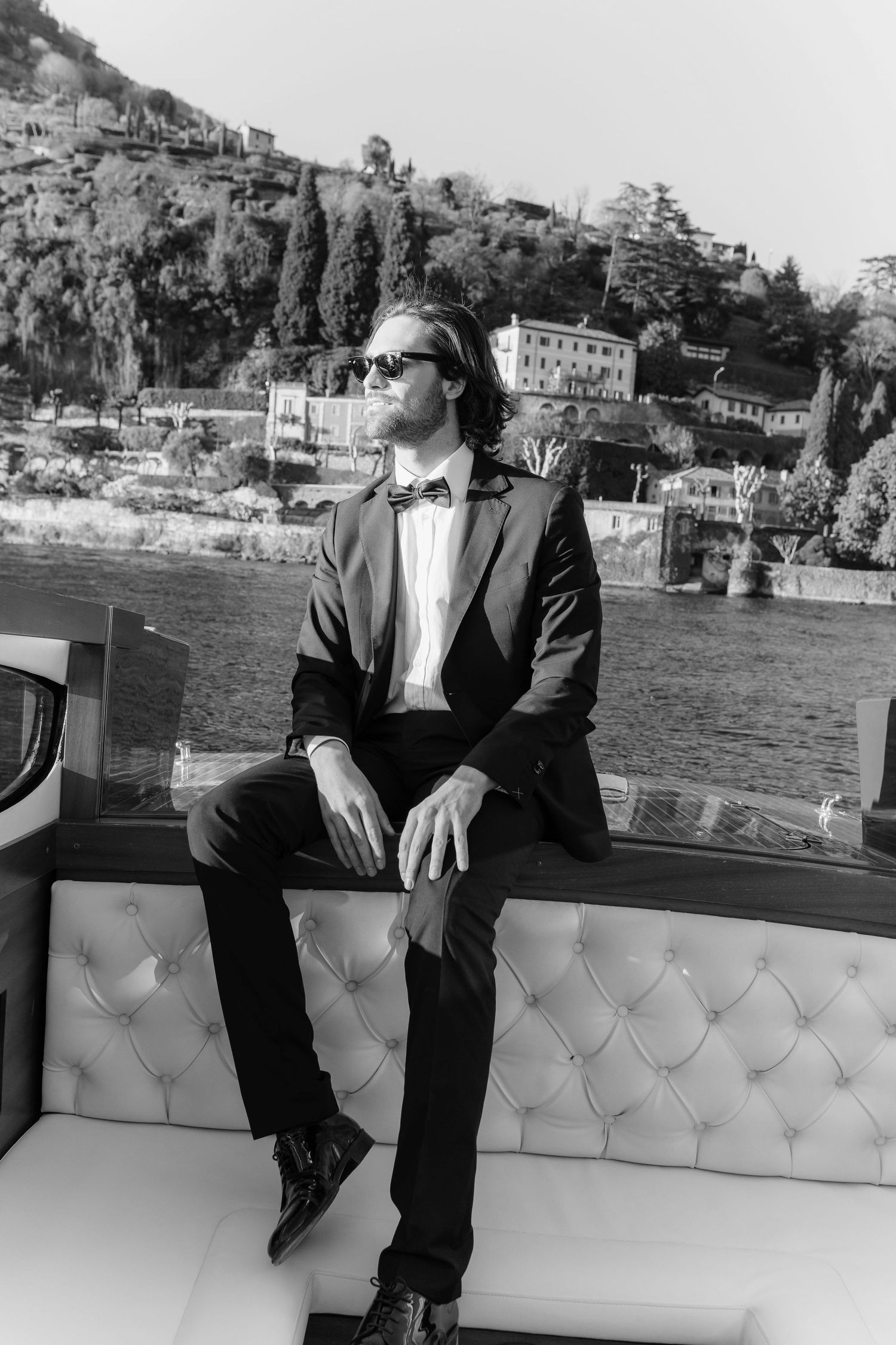 Black and white photo of groom in tuxedo and sunglasses sitting on a boat at Lake Como, Italy.
