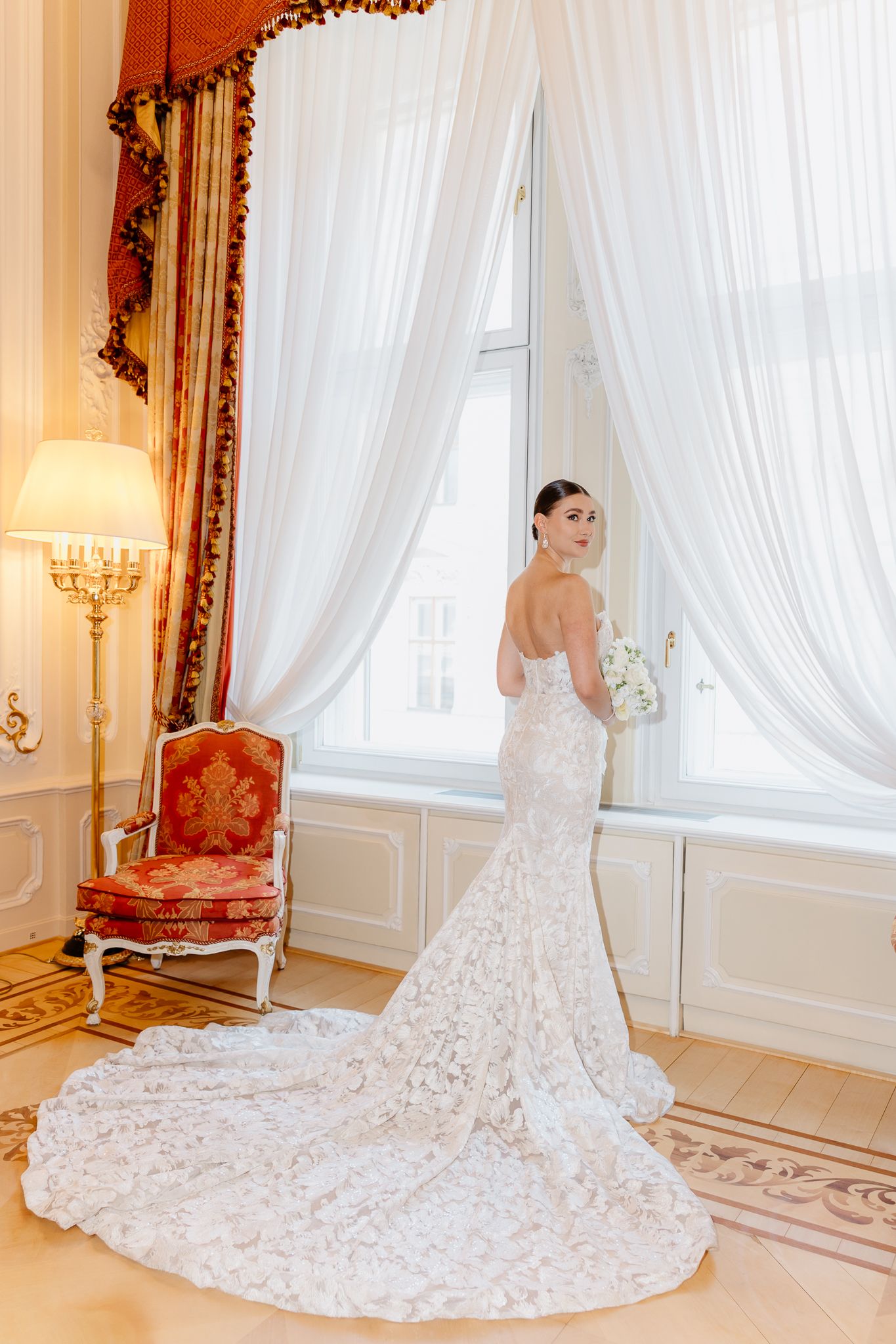 Bride in fitted lace mermaid gown with long train standing at window holding white bouquet in Hotel Imperial,Vienna.
