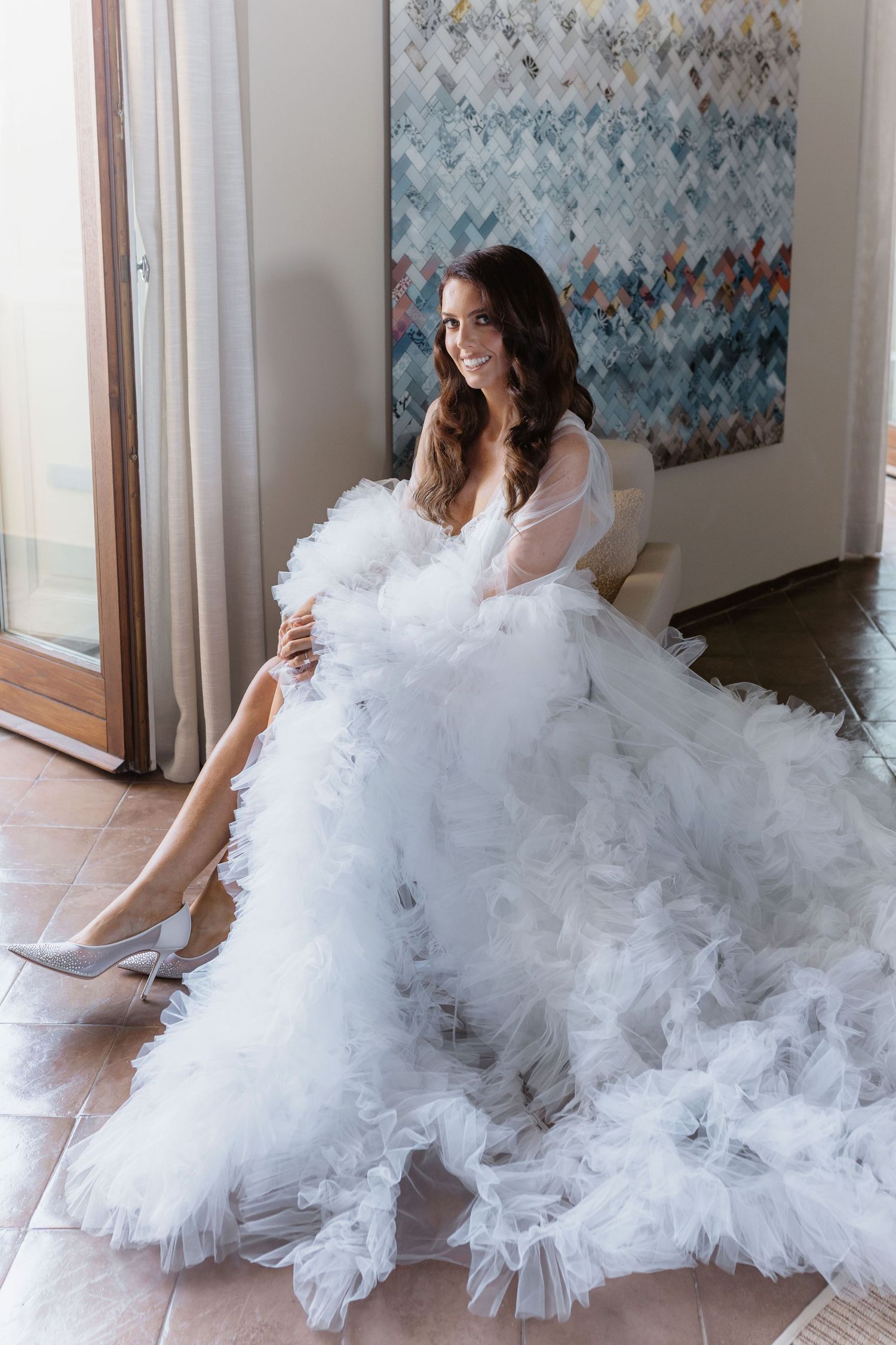 Getting ready bride in a voluminous white tulle wedding gown with sheer sleeves sits elegantly in a sunlit room in Italy.