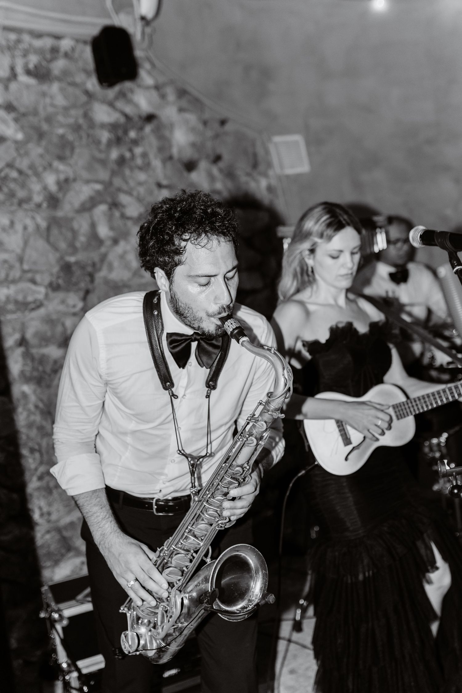 Black and white photo of a man playing saxophone with a female guitarist performing live at an elegant event.