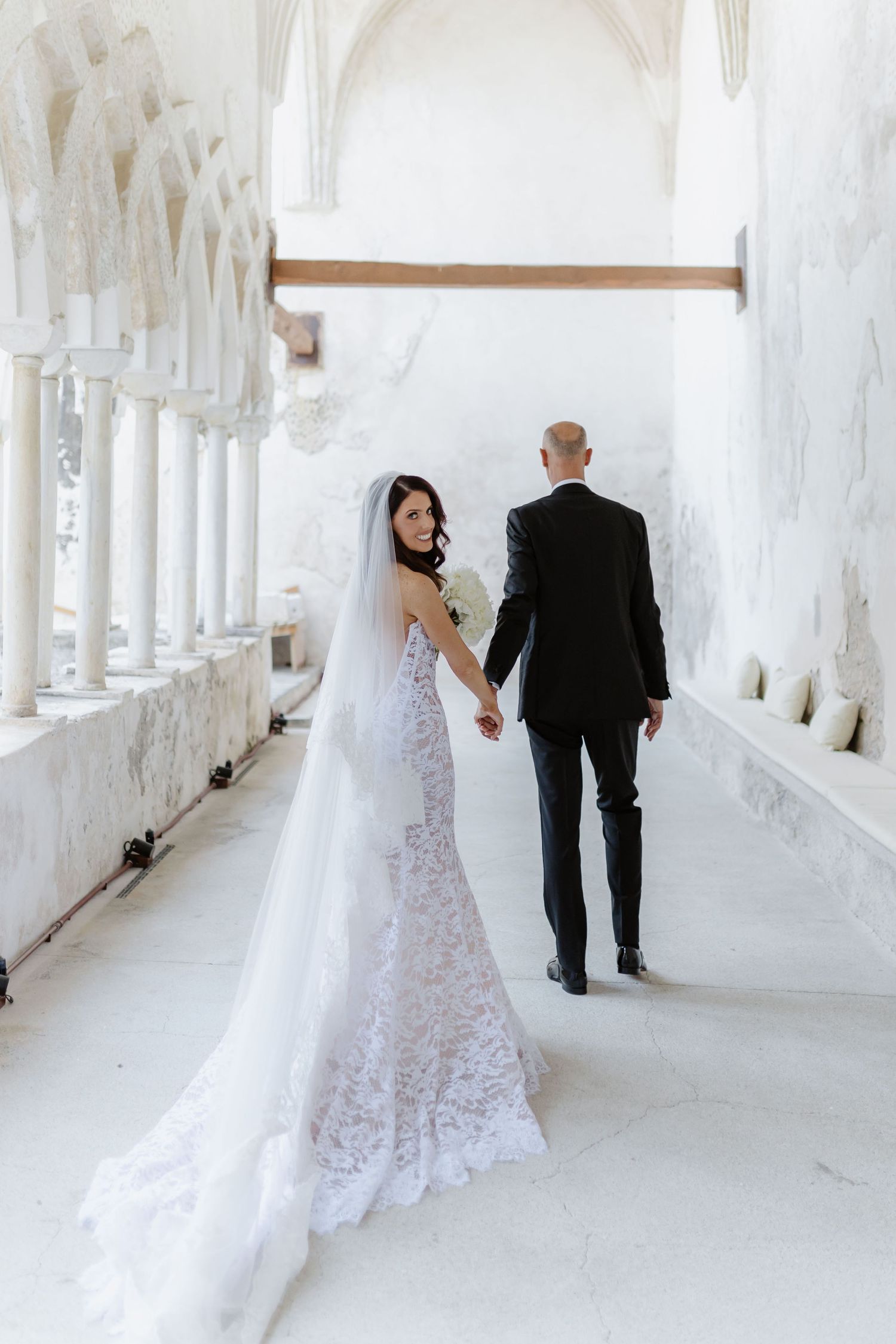 Bride in lace gown and veil holds groom's hand walking through white marble corridor.