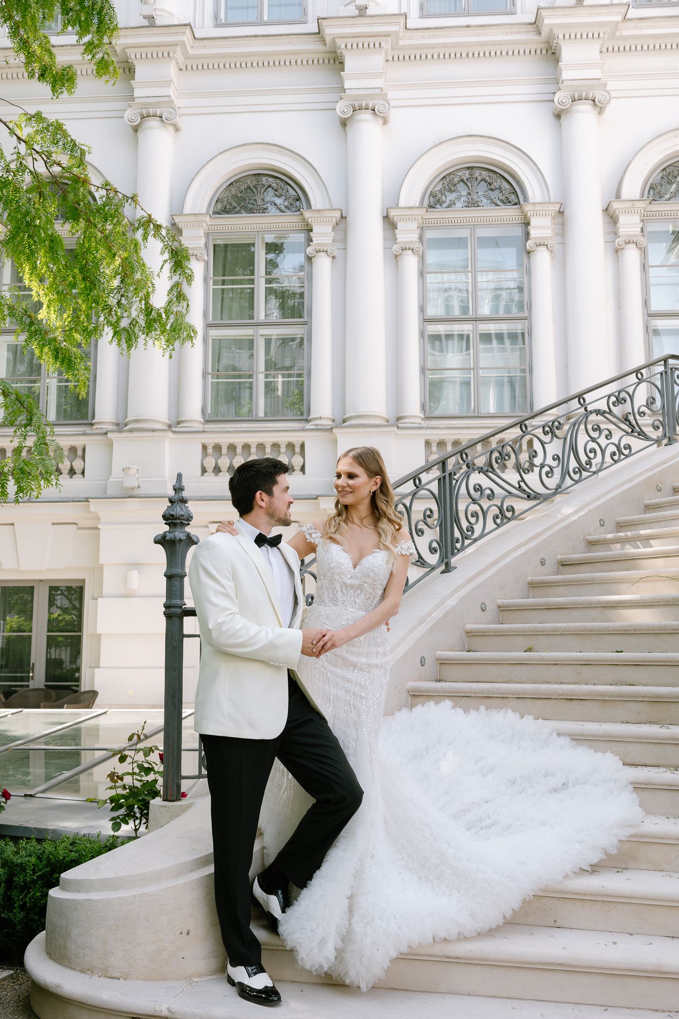 Bride in white lace gown and groom in white tuxedo pose on ornate staircase outside elegant neoclassical building.