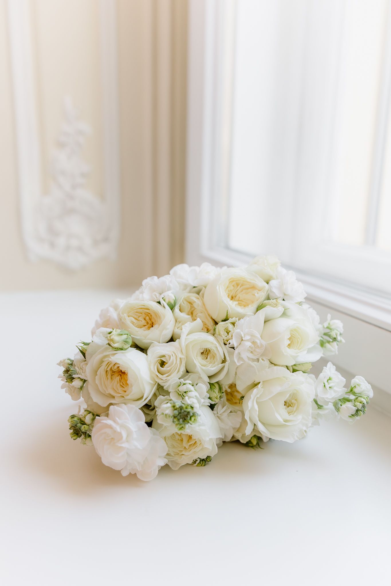 Luxury bridal bouquet of white garden roses and ranunculus resting on white surface near window.