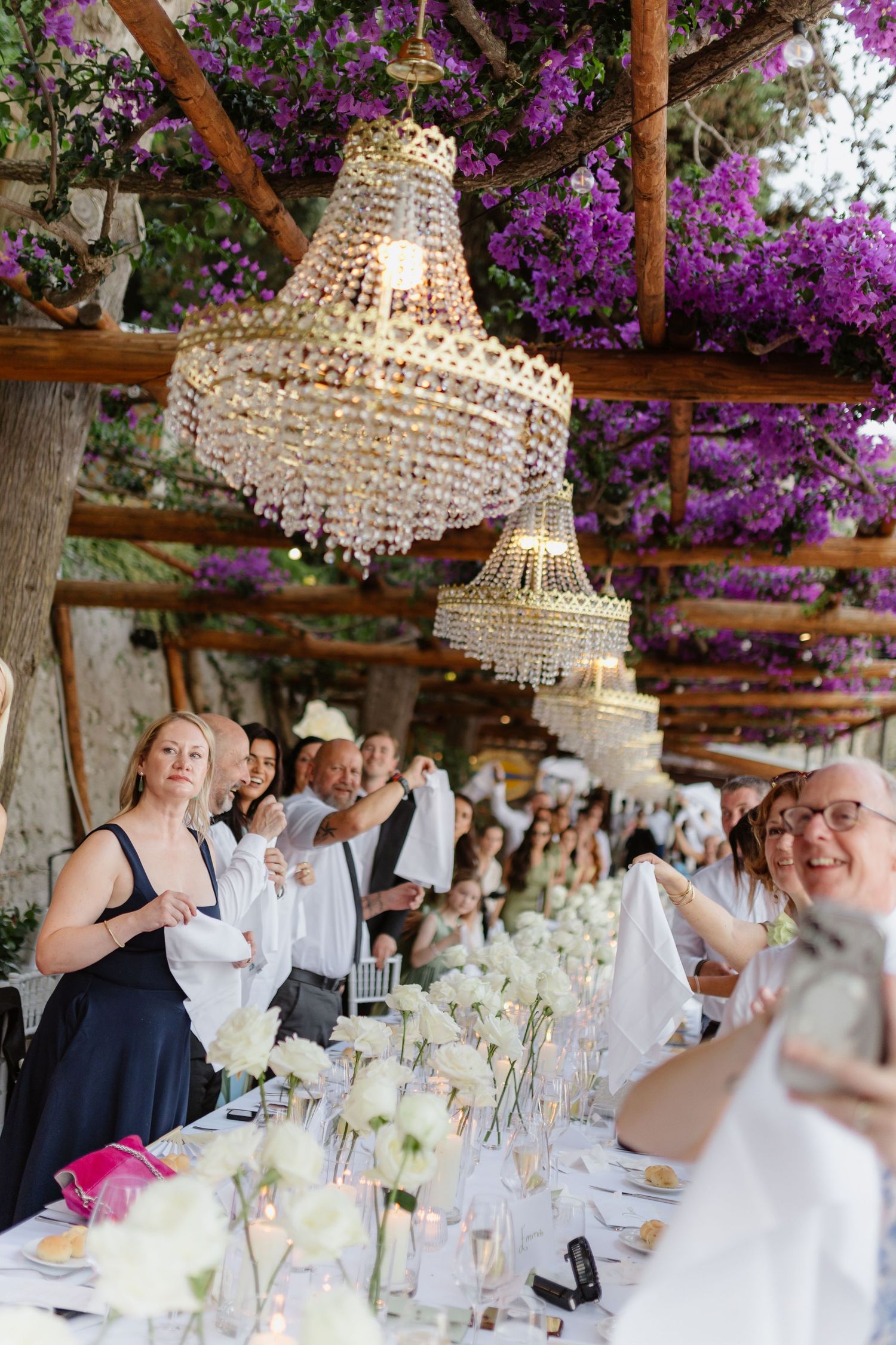 Elegant outdoor wedding reception with crystal chandeliers, white floral centerpieces, and purple bougainvillea overhead.