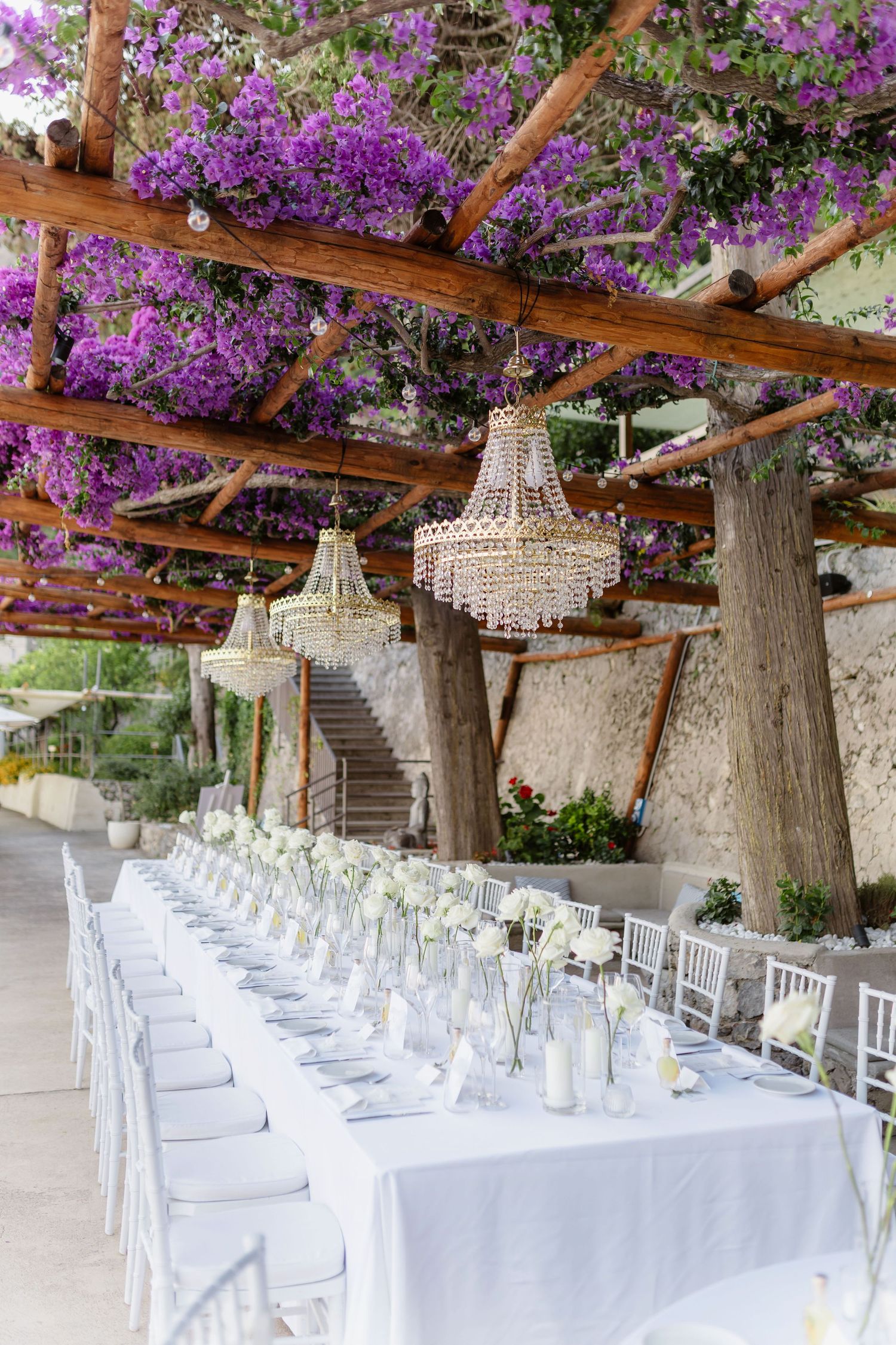 Elegant outdoor wedding reception table set under a pergola with purple bougainvillea and crystal chandeliers.
