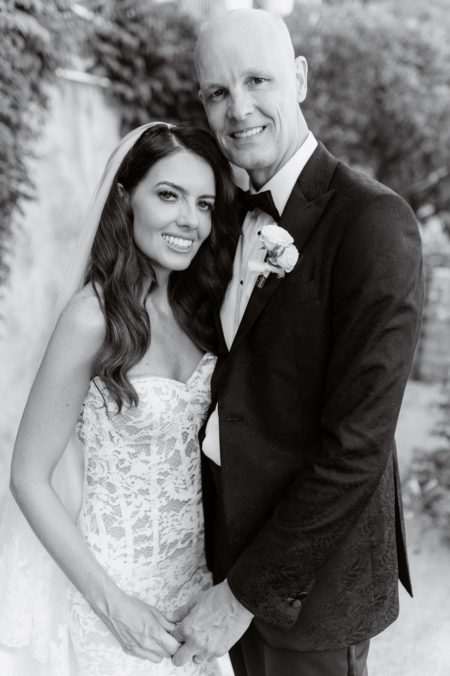 Black and white wedding portrait of smiling bride in lace gown and groom in tuxedo with boutonniere.