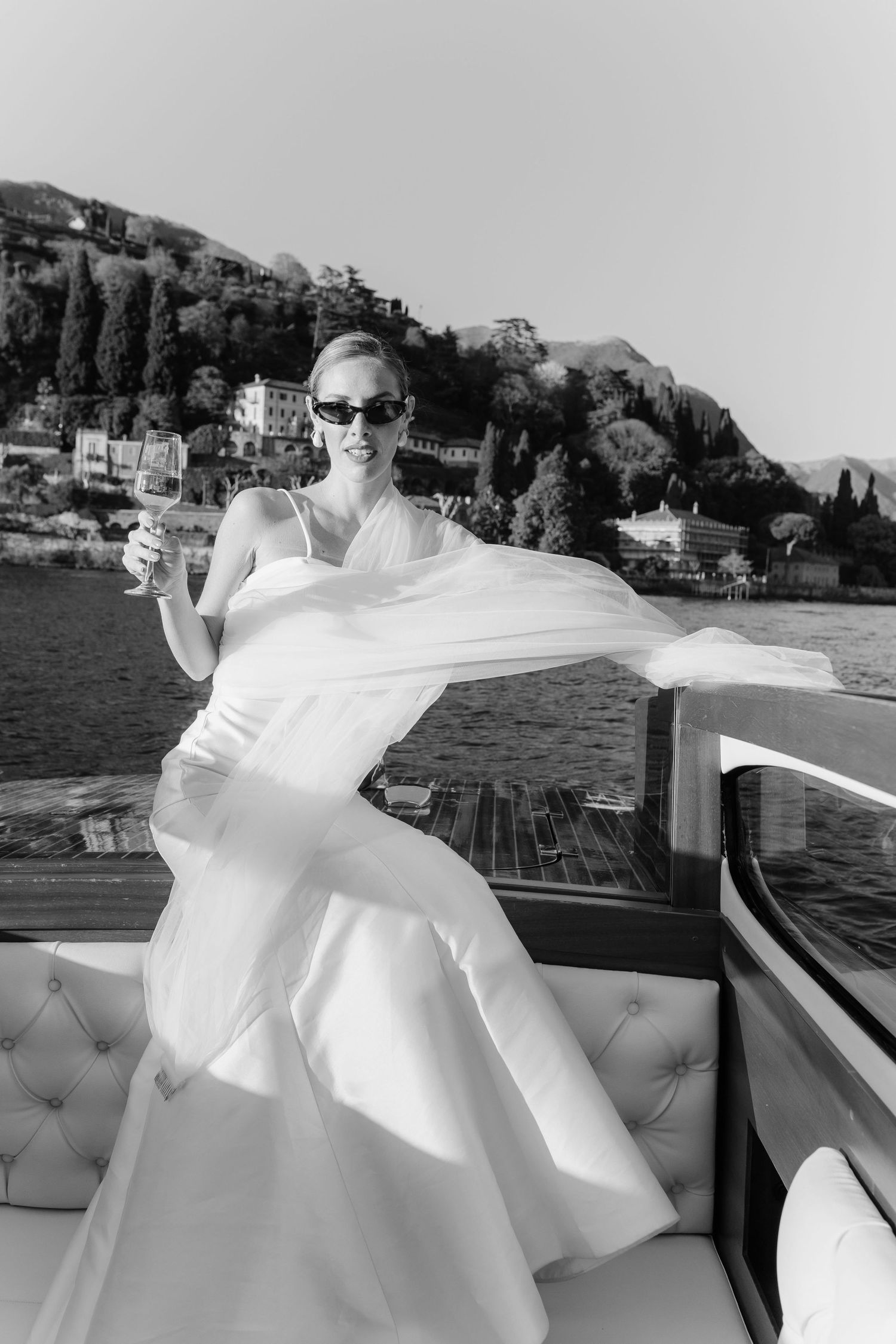 Black and white photo of a bride in a flowing white gown holding a wine glass on a luxury boat with scenic hills behind.