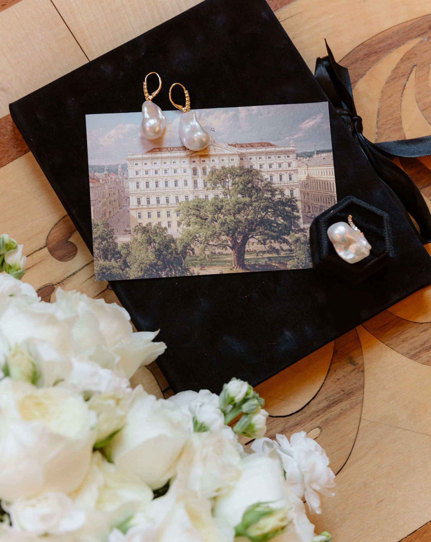 Gold baroque pearl drop earrings displayed on black velvet beside a hotel postcard and white flowers.