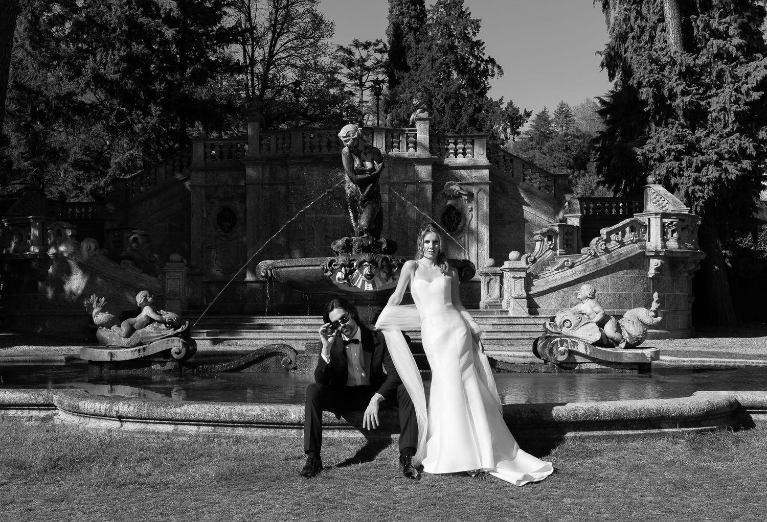 Dramatic black and white wedding photo of couple posing before an ornate garden fountain sculpture.