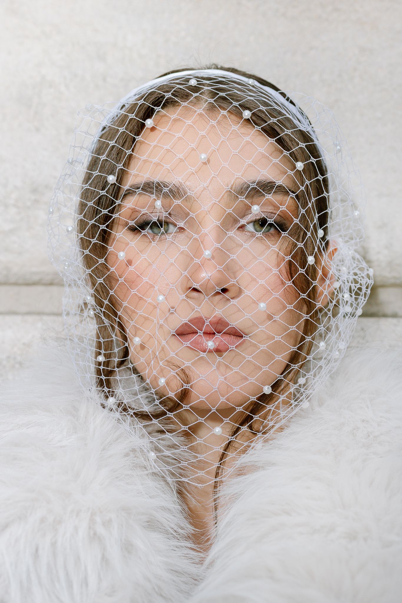 Close portrait of bride wearing pearl-embellished birdcage veil with white fur wrap.