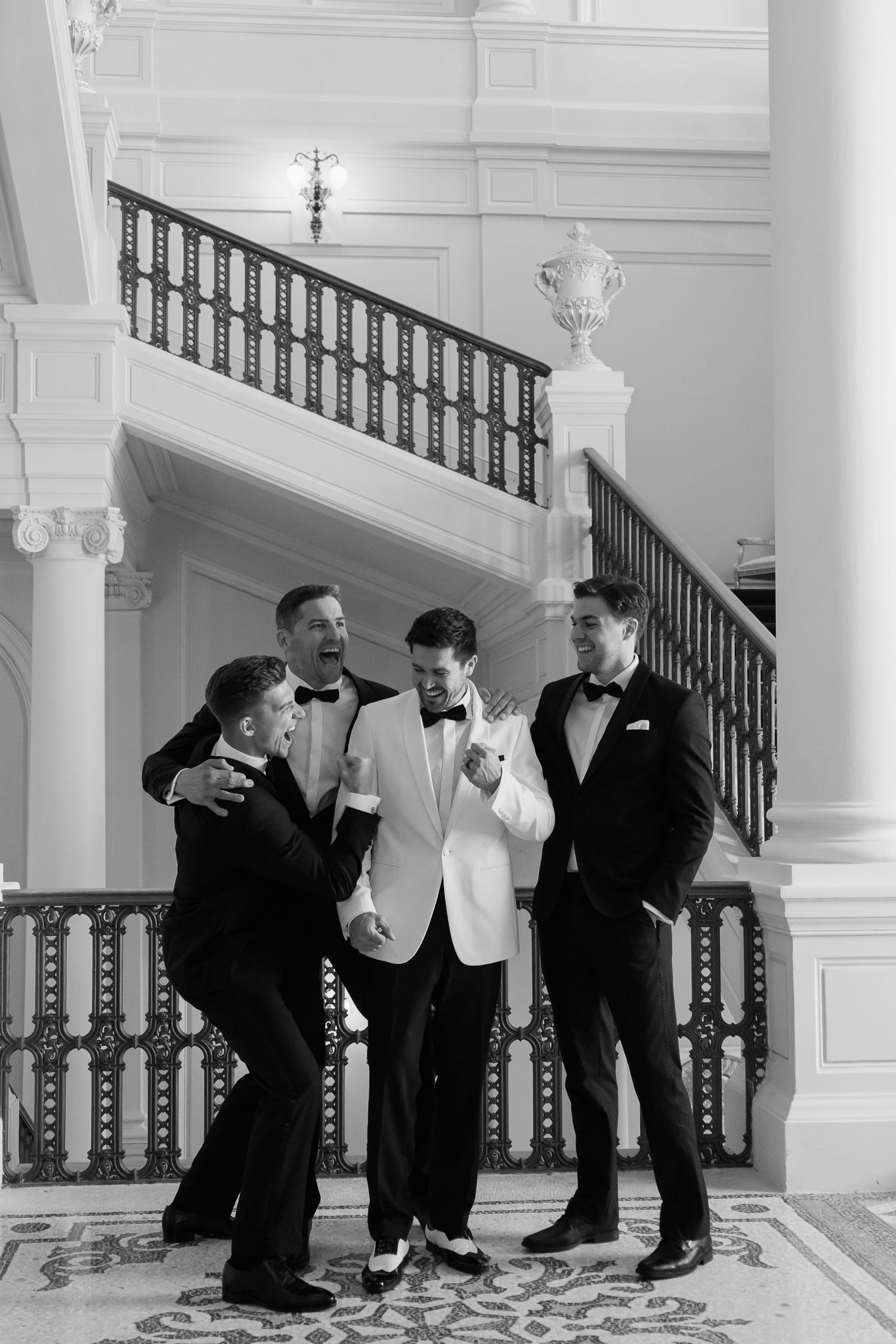 Black and white photo of groom in white tuxedo laughing with groomsmen on elegant staircase.