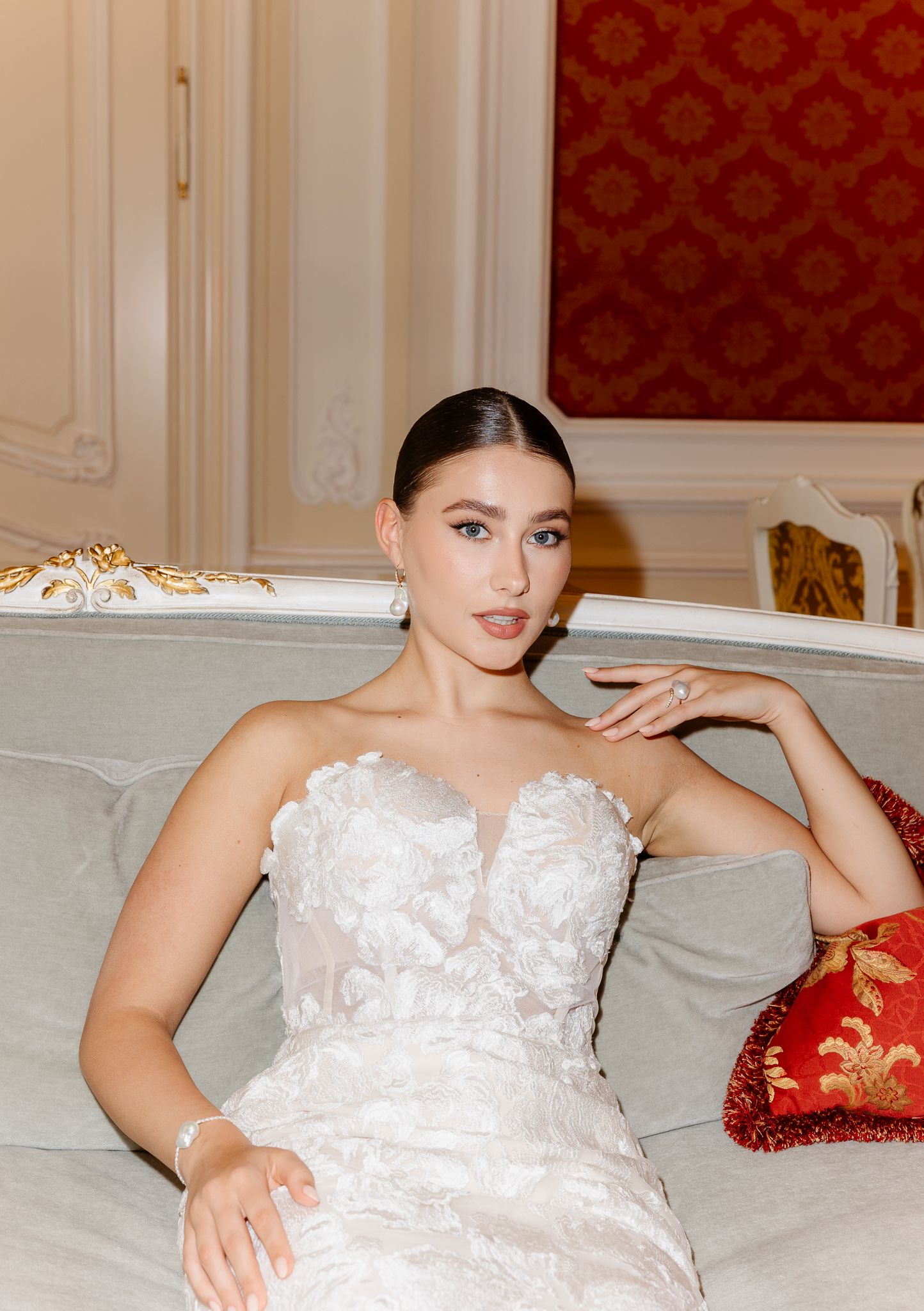 Beautiful bride in a floral applique strapless gown relaxes on a sofa in a opulent red room.