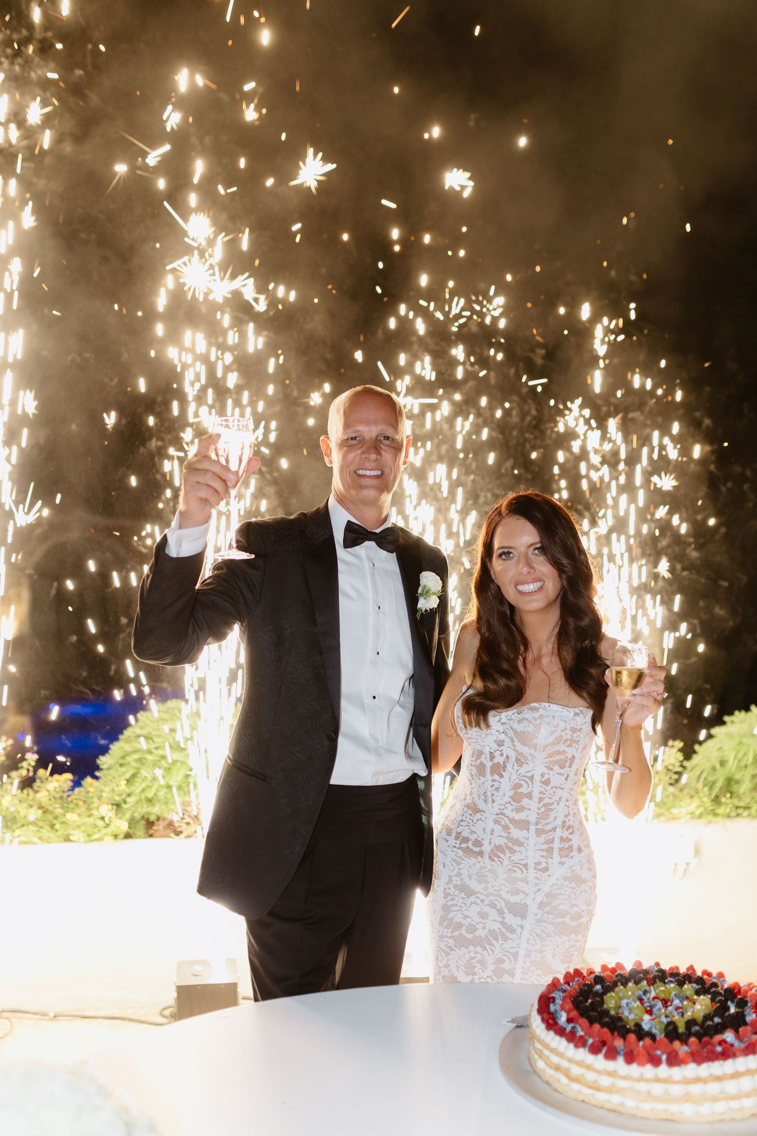 Bride and groom toasting champagne with fireworks backdrop beside a fruit-topped wedding cake.