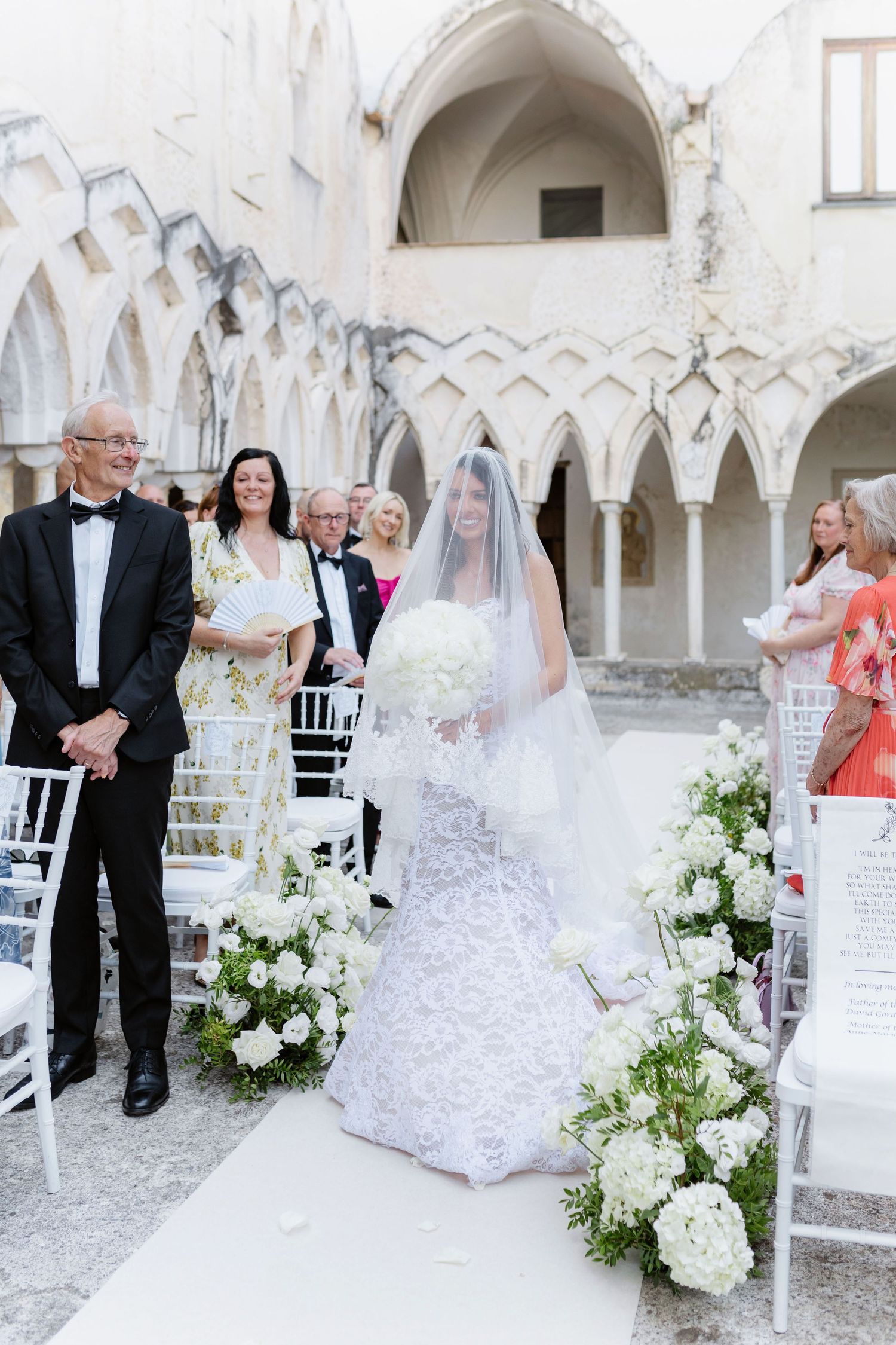 Bride in lace gown and veil walks down outdoor aisle with white floral arrangements at elegant wedding ceremony.