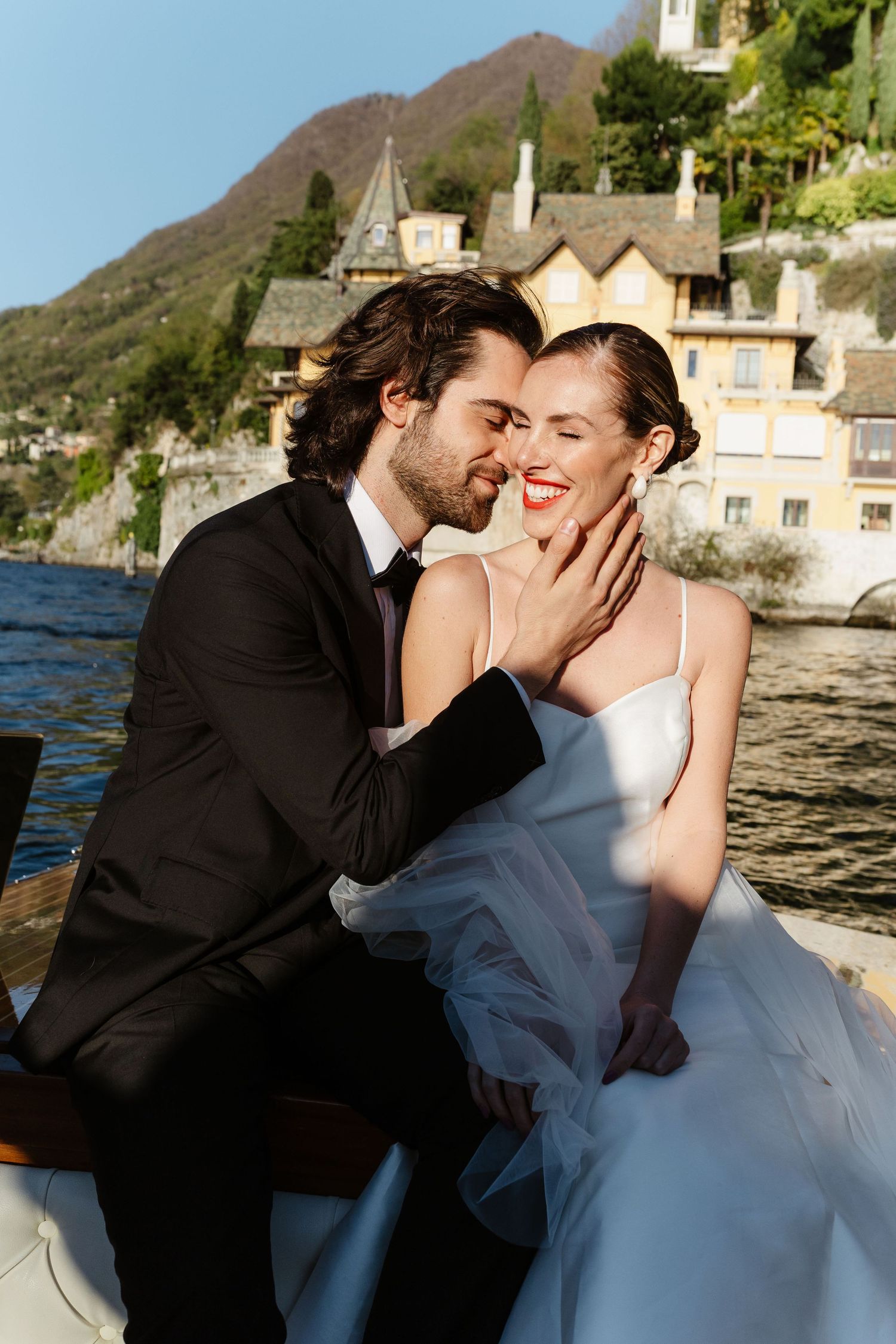 Groom gently holding bride's face near a stunning yellow lakeside villa on a bright sunny day.