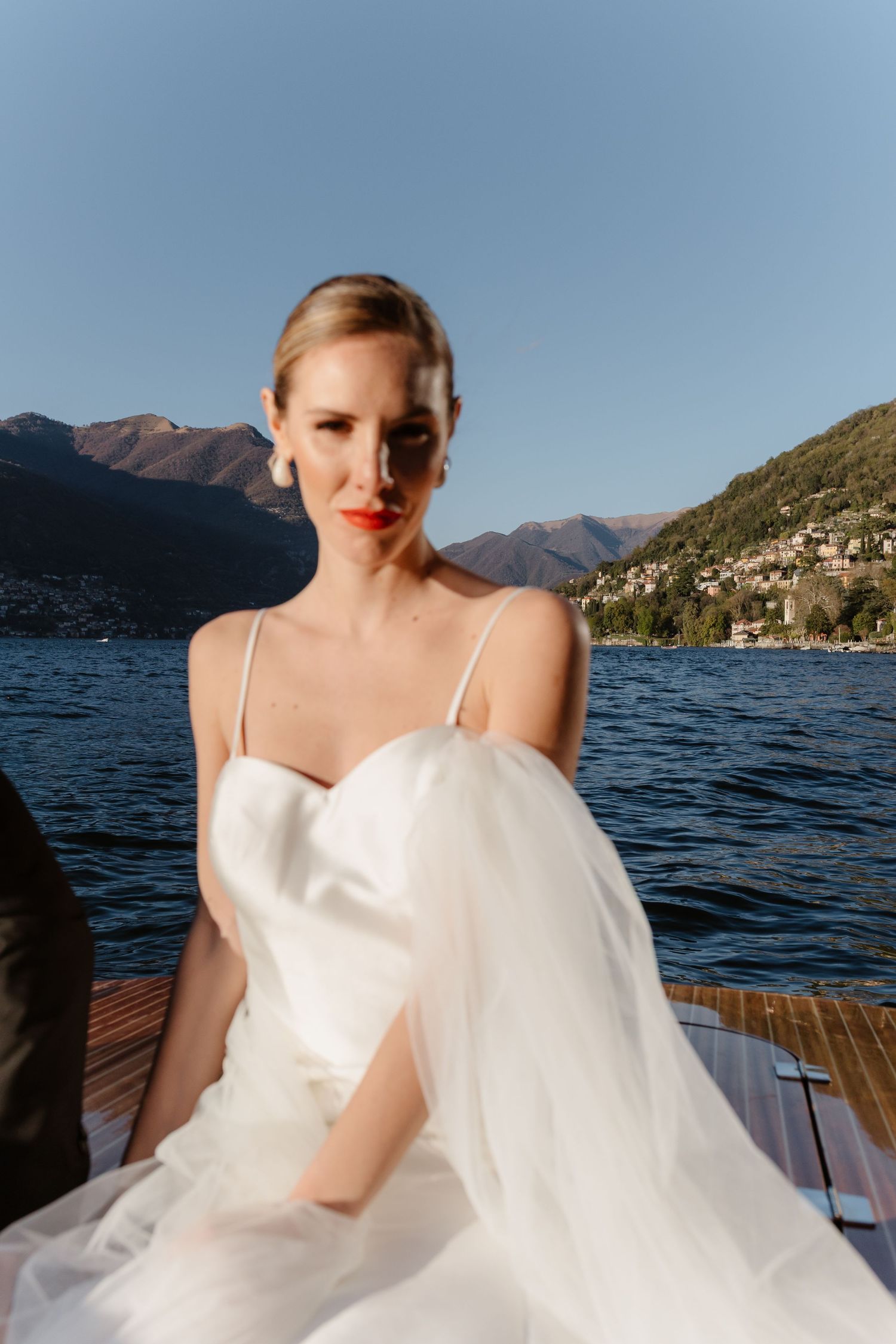 Bride in white spaghetti strap gown poses on boat with blue mountain lake behind her.