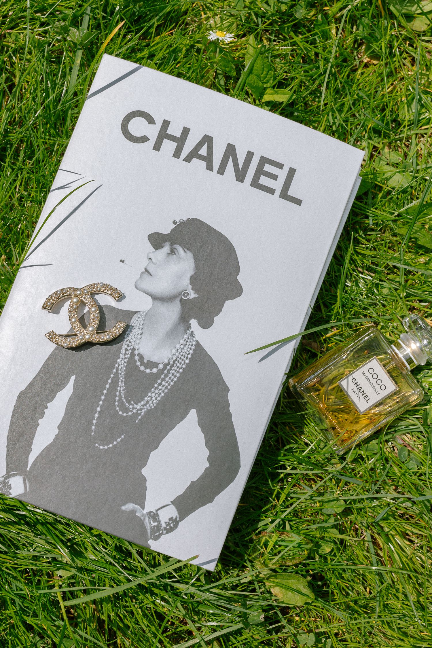 Chanel book with CC brooch and Coco Chanel perfume bottle resting on lush green grass.