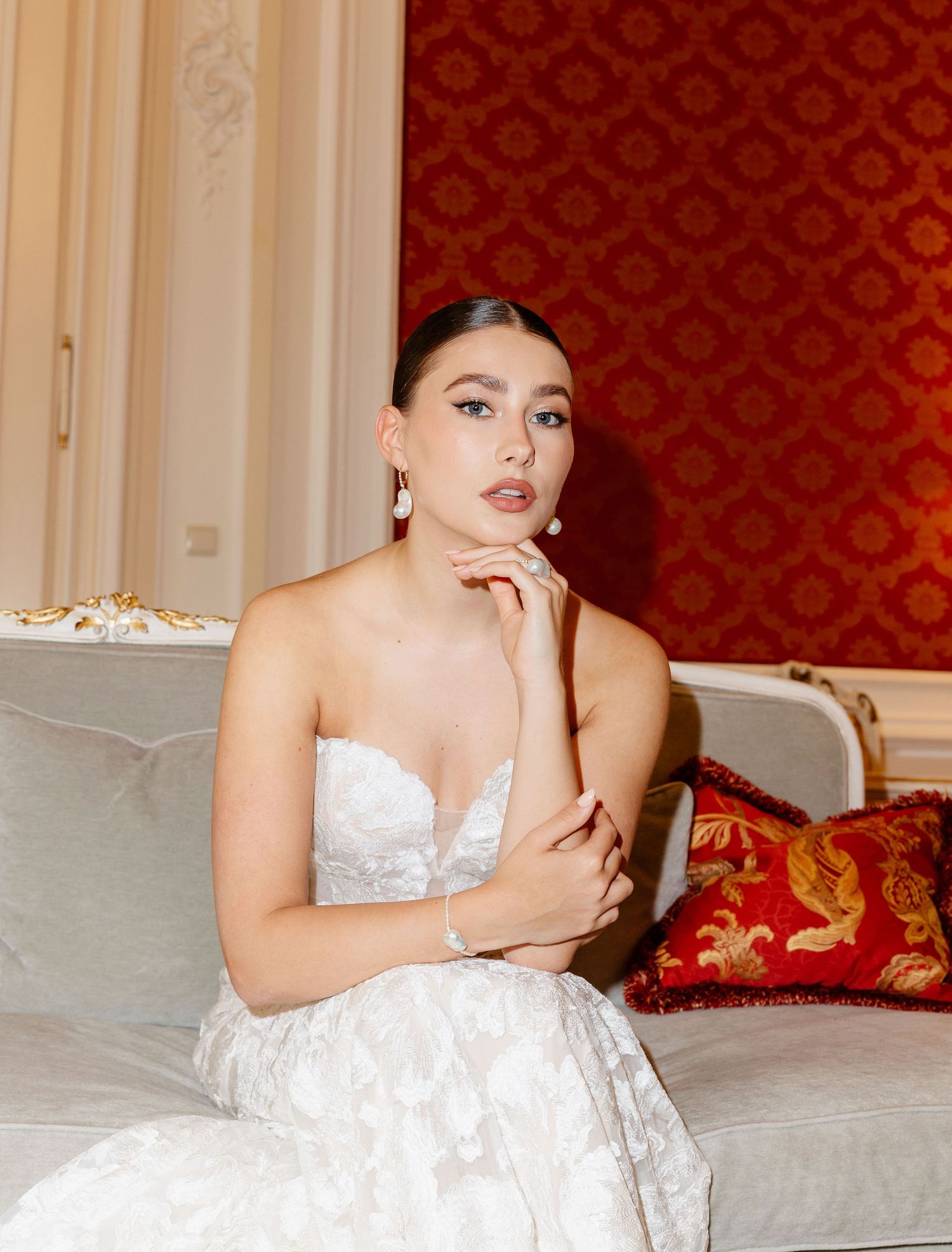 Beautiful bride in strapless lace wedding dress seated on sofa against rich red wallpaper background.