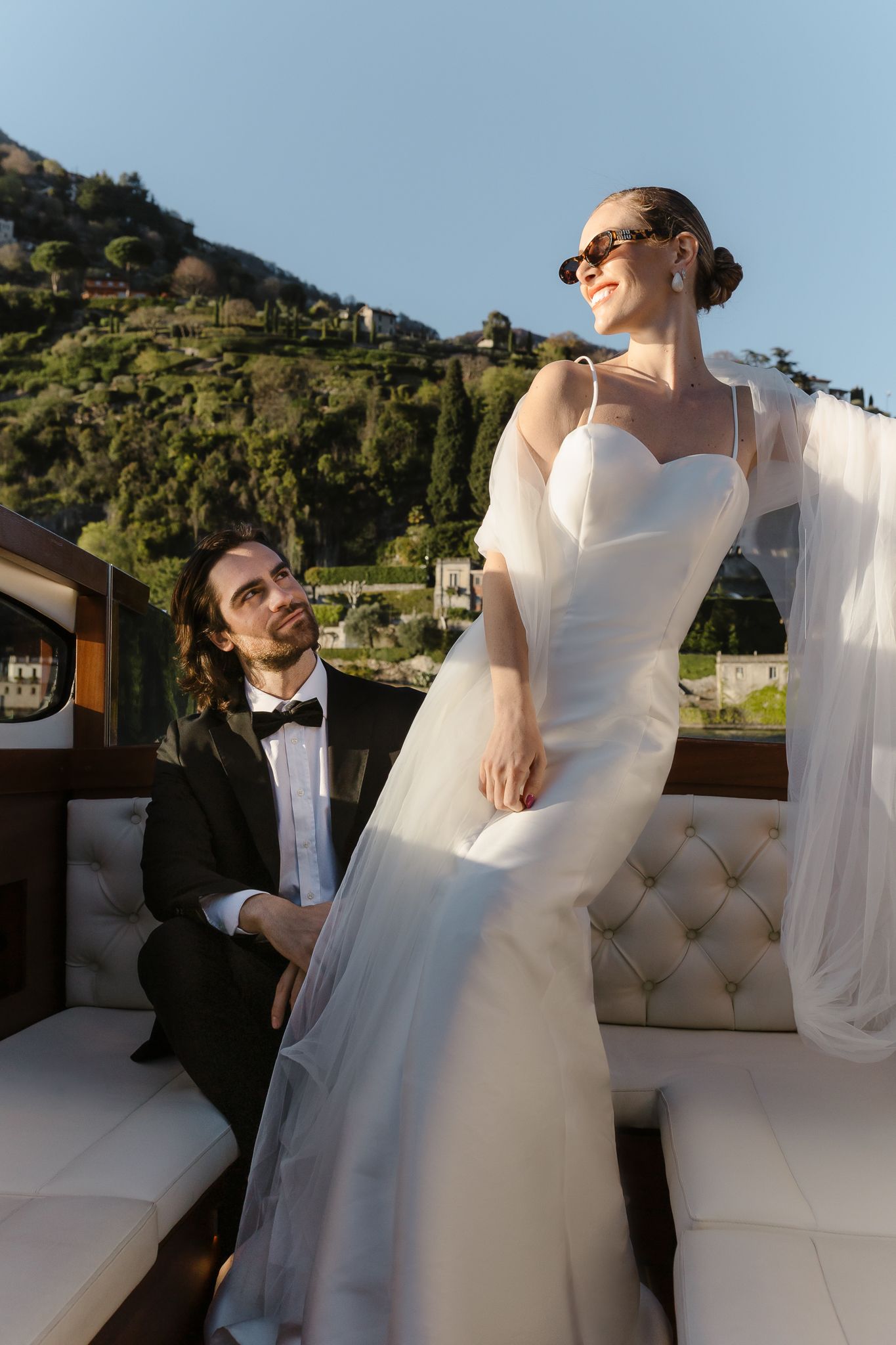 Bride in sunglasses and white gown stands on luxury boat as groom looks up adoringly.