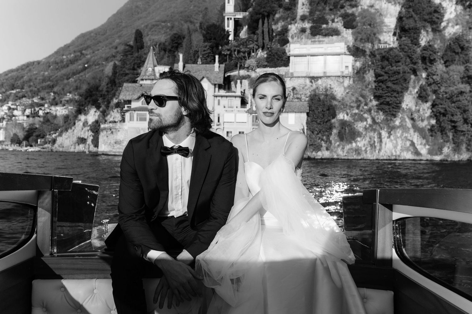Black and white photo of bride and groom sitting on a boat with scenic lakeside architecture behind.