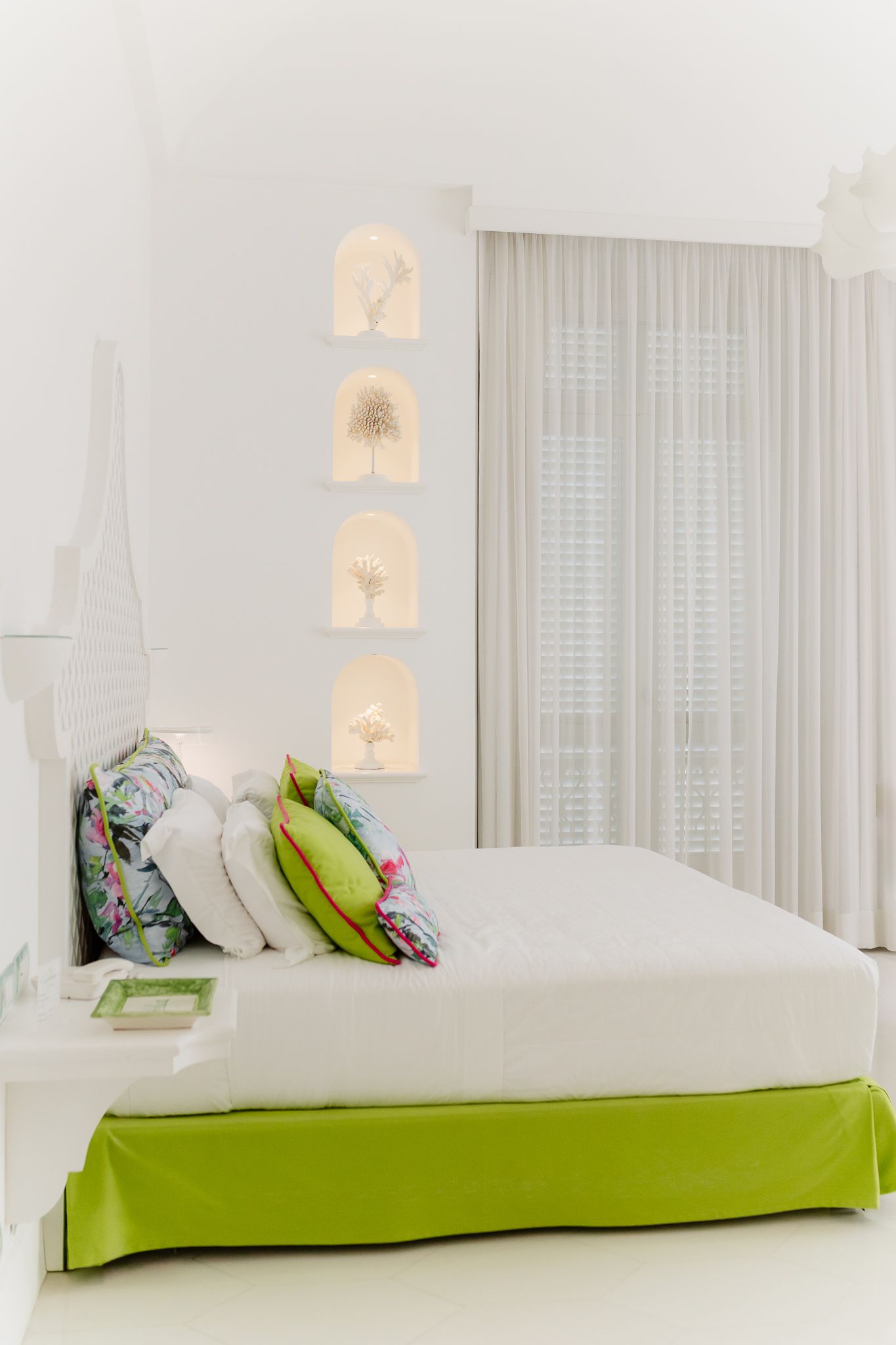 Elegant white bedroom with green bed skirt, floral pillows, illuminated wall niches, and sheer curtains.