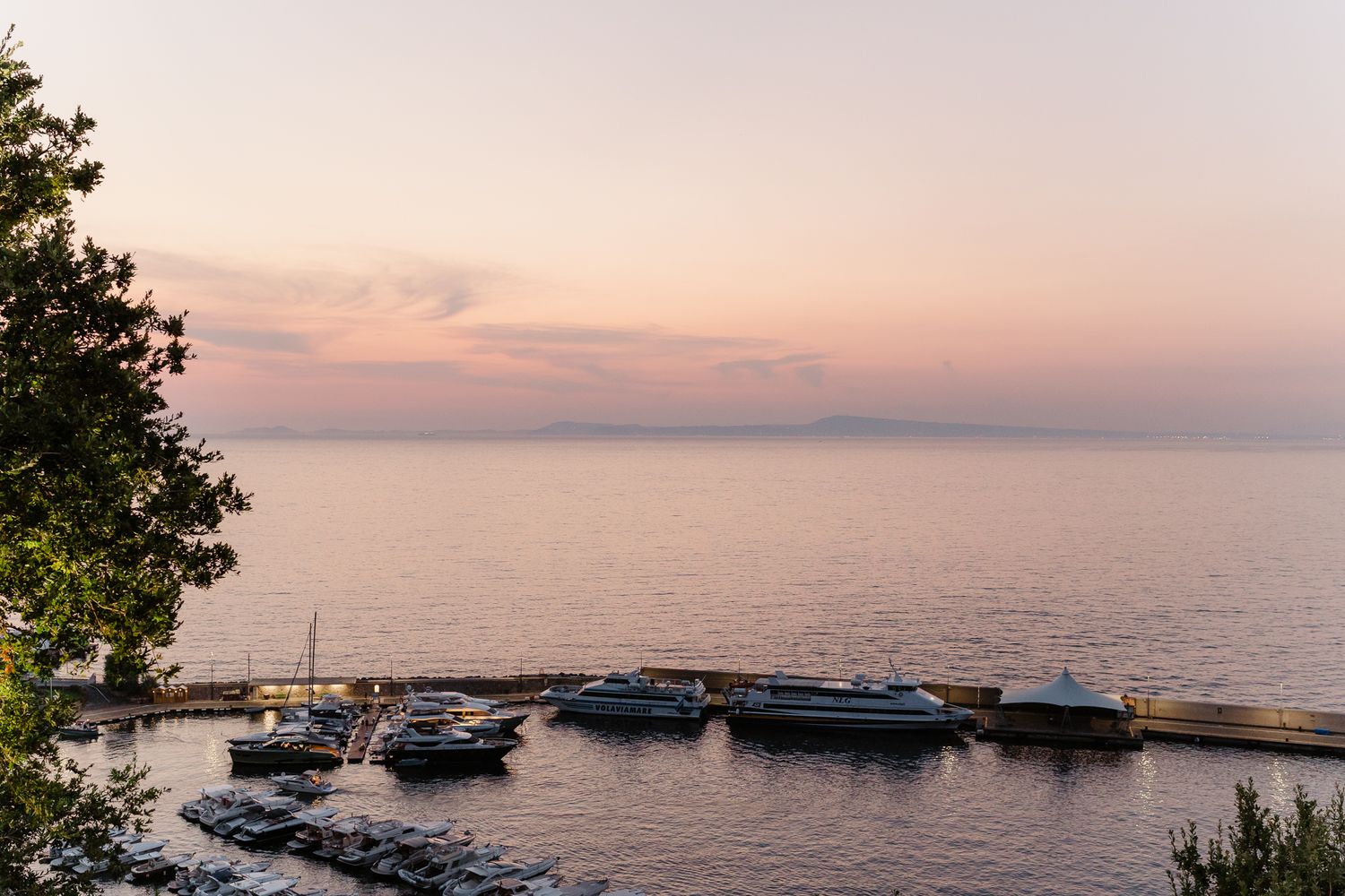 Serene sunset over calm ocean with boats docked at a coastal marina.