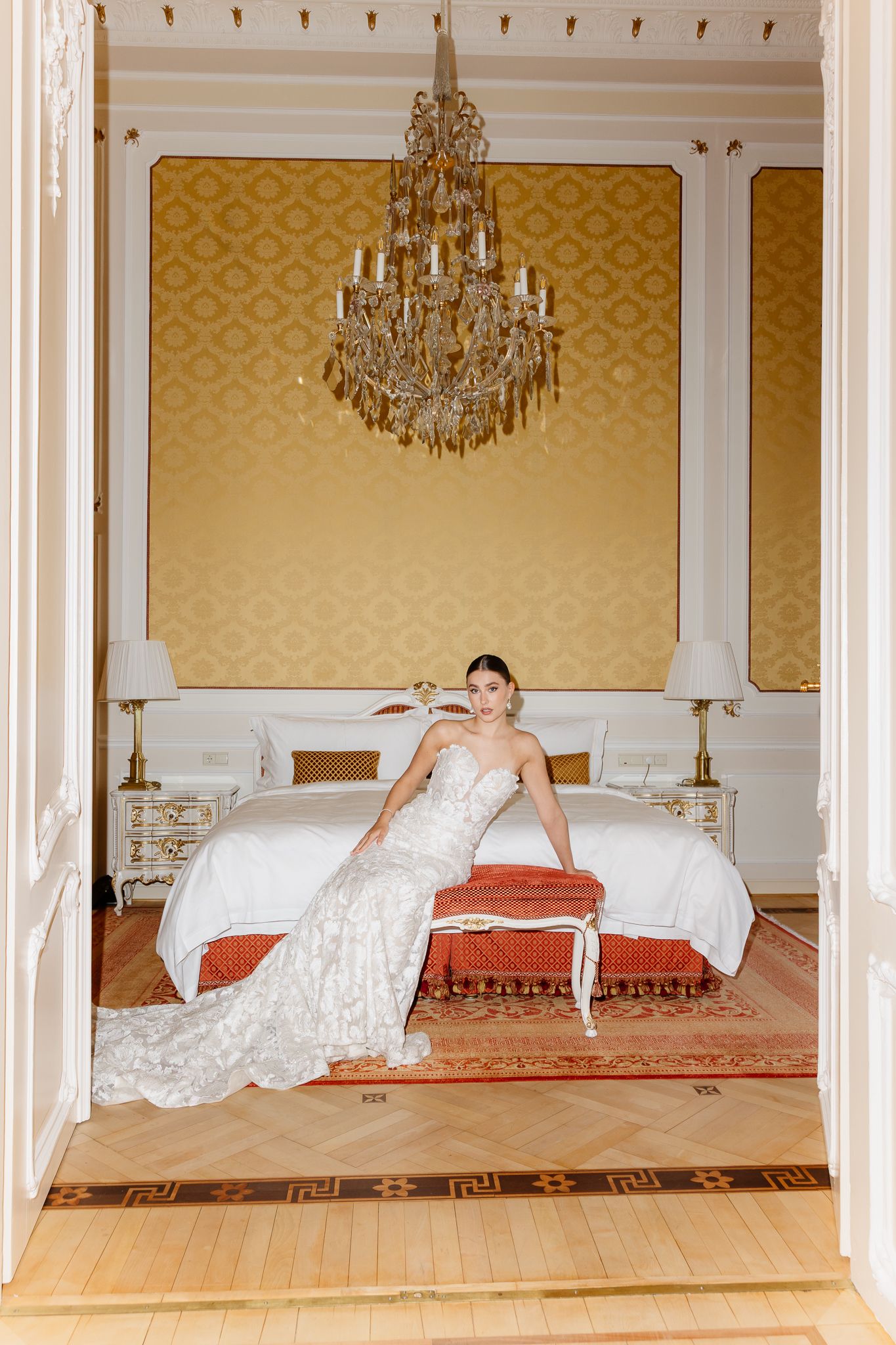 Bride in lace mermaid wedding gown sits on bed bench in golden wallpapered room with crystal chandelier.