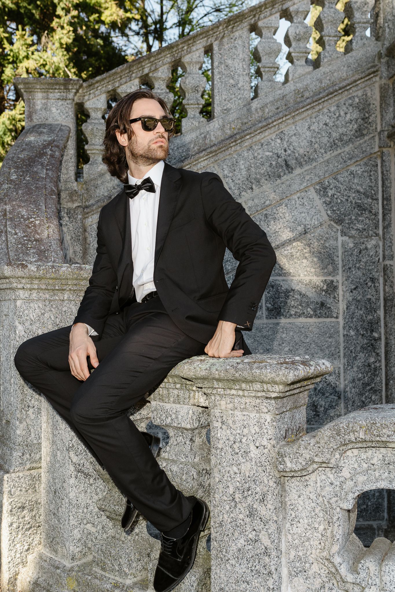 Stylish groom in black tuxedo and sunglasses seated on stone balustrade at a classic villa.