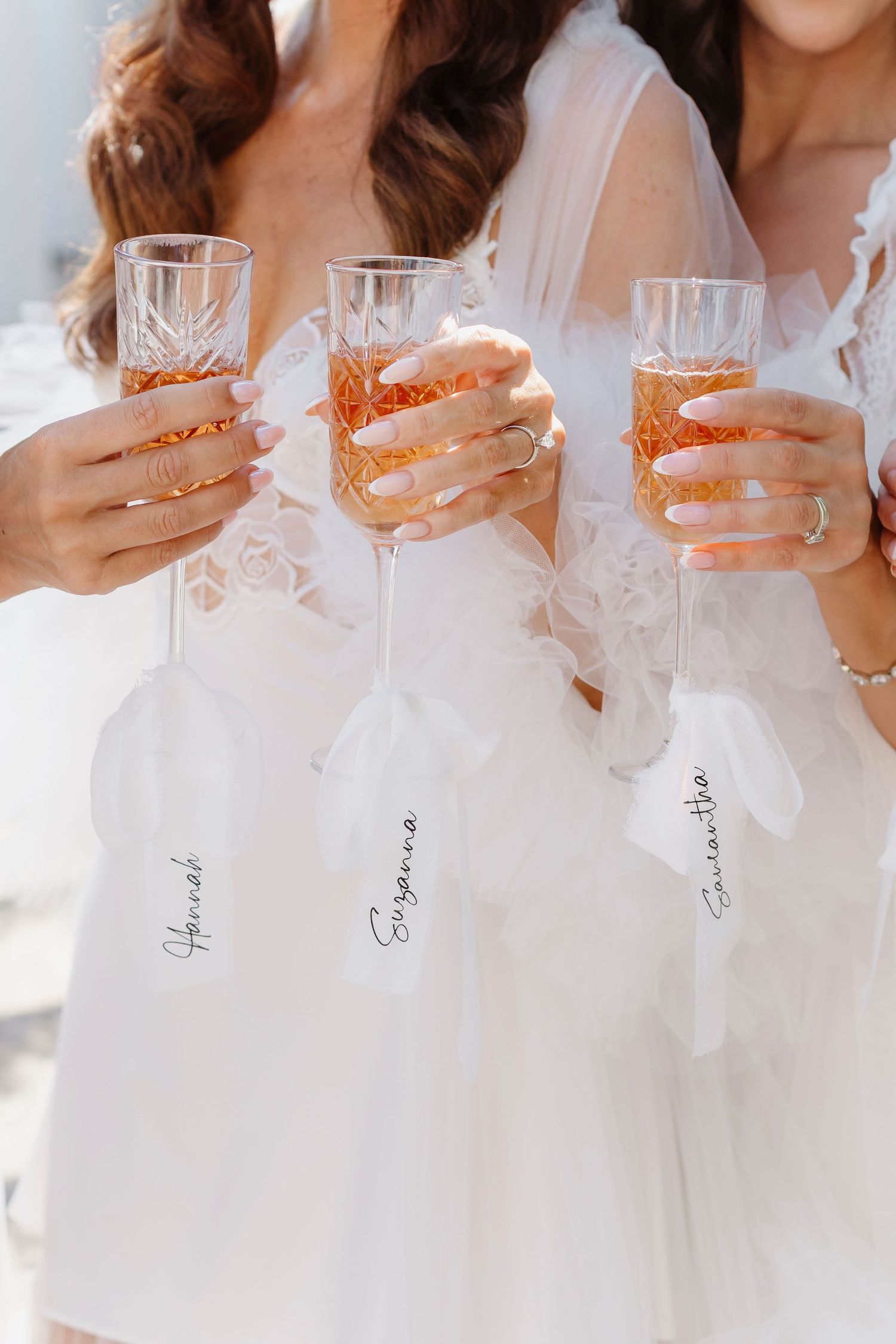 Three bridesmaids in white robes holding personalized champagne flutes with rose champagne at a bridal celebration.