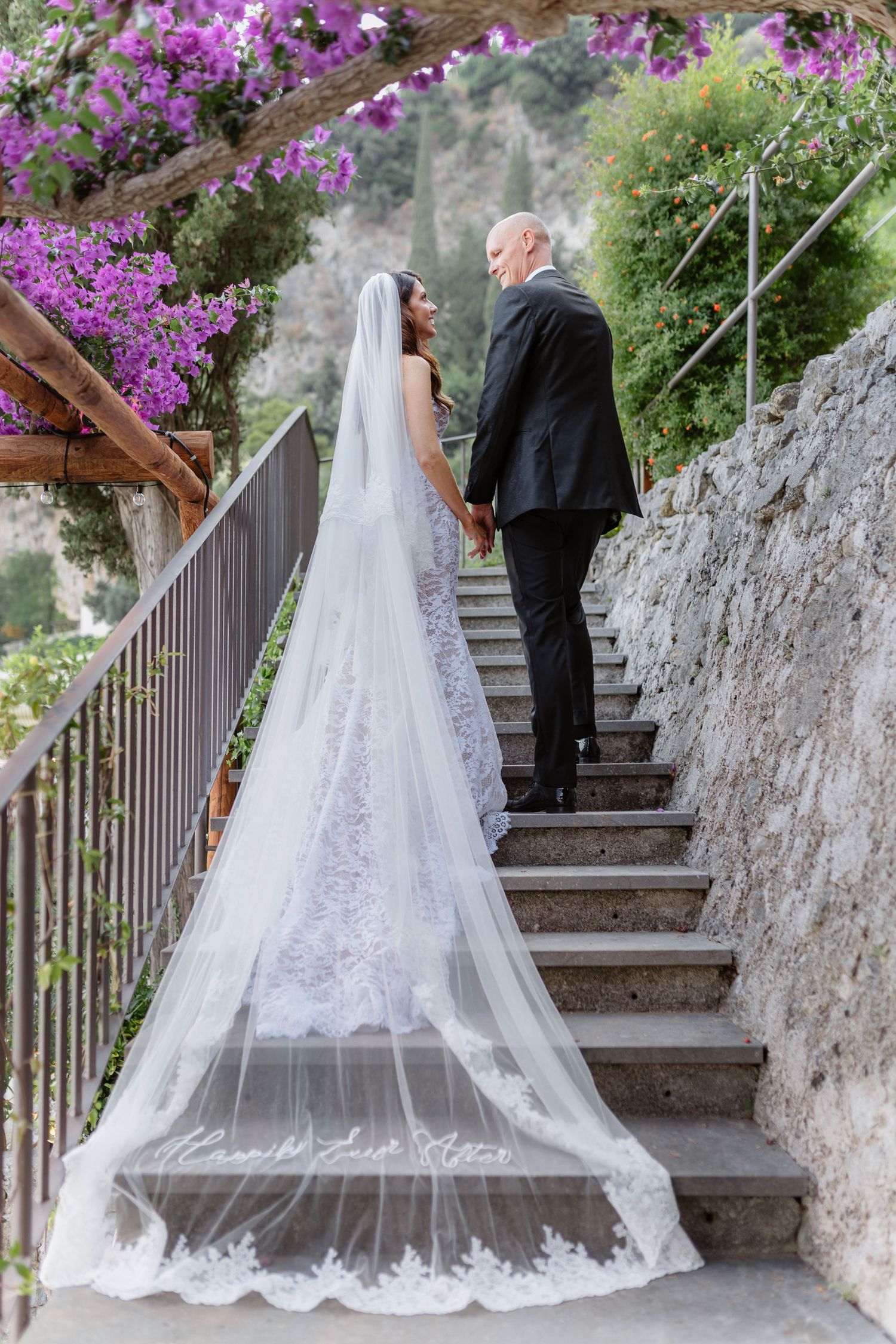 Bride in lace gown with cathedral veil walks stone steps with groom amid pink bougainvillea at scenic wedding venue.
