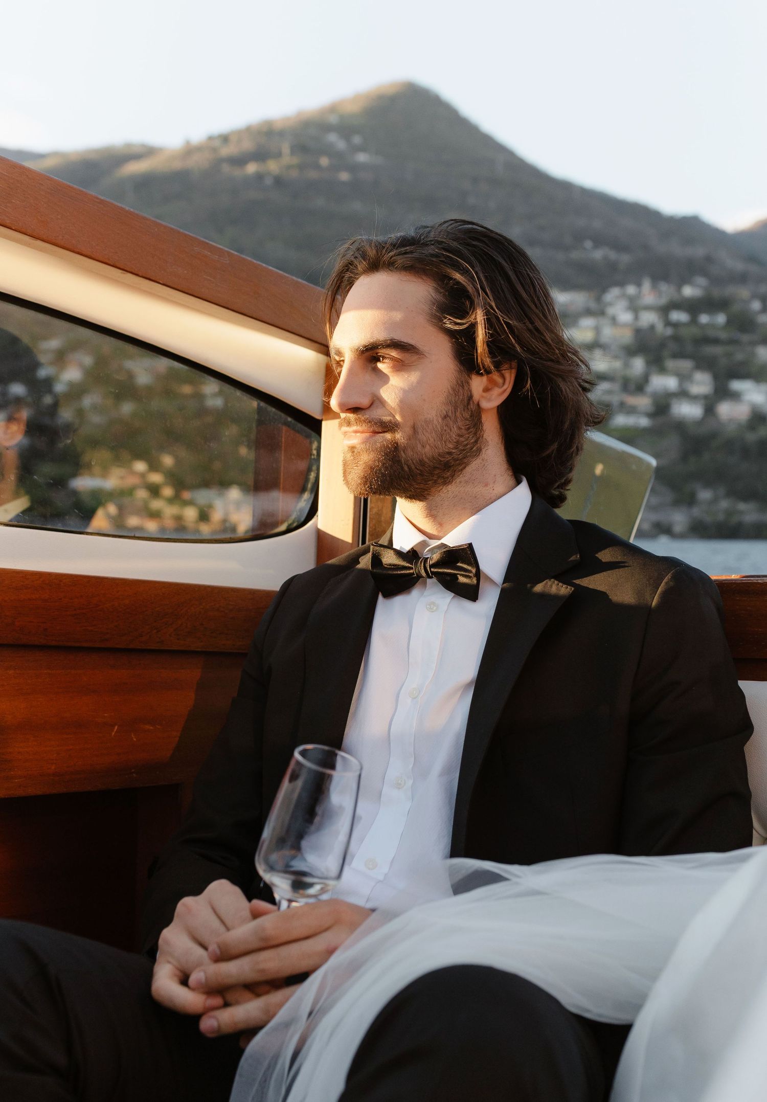 Handsome groom in black tuxedo holding champagne glass on luxury yacht with golden sunset and mountain view.
