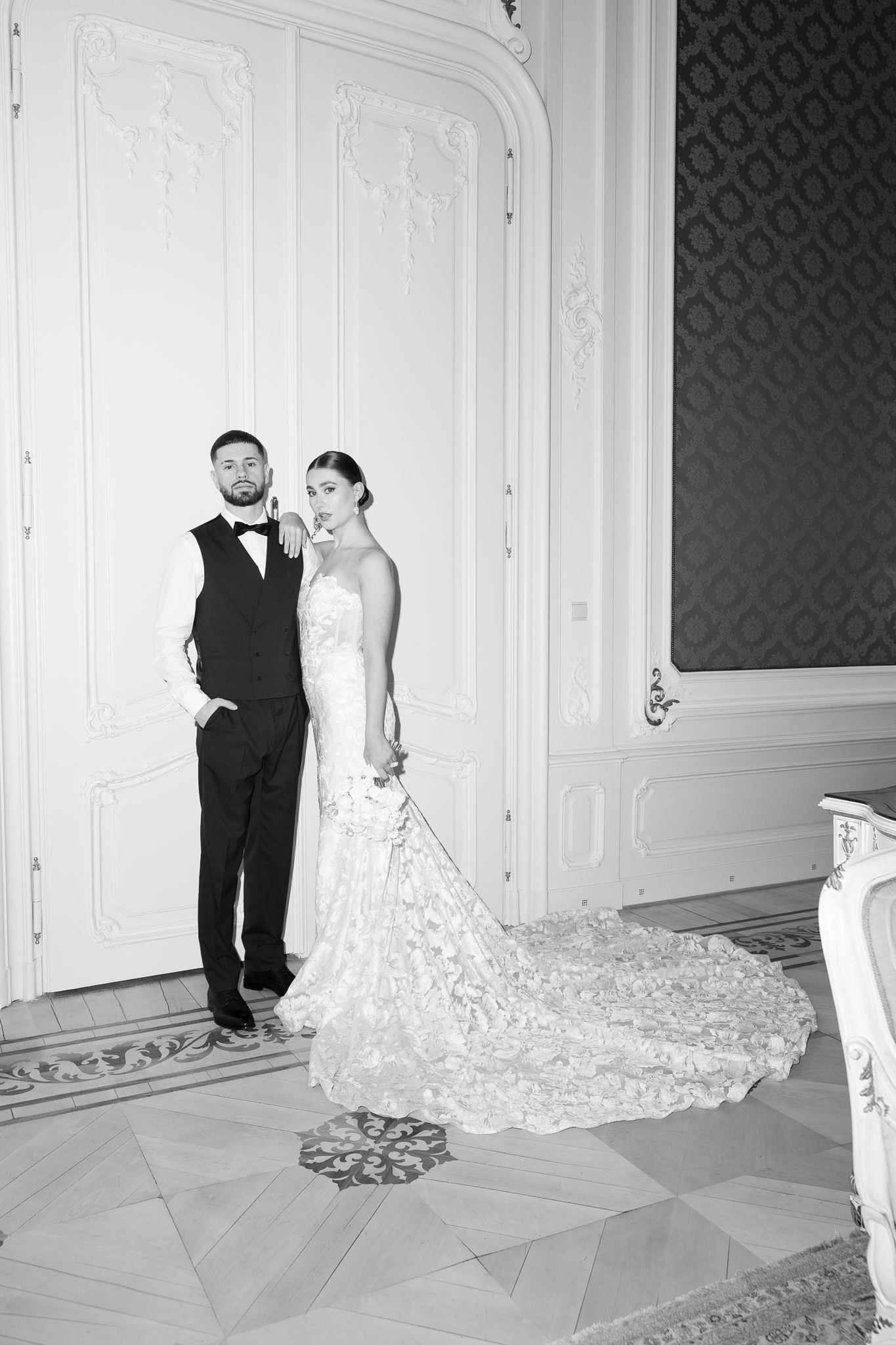 Black and white portrait of bride and groom posing elegantly in an ornate ballroom interior.