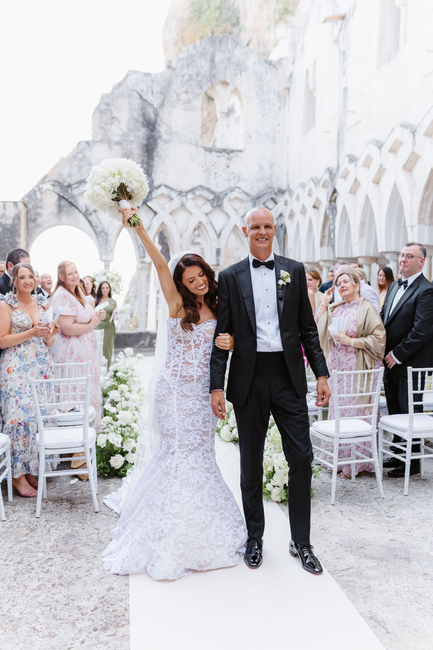 Bride raises bouquet triumphantly while walking down aisle with groom in tuxedo at elegant outdoor wedding ceremony in Amalfi Coast.