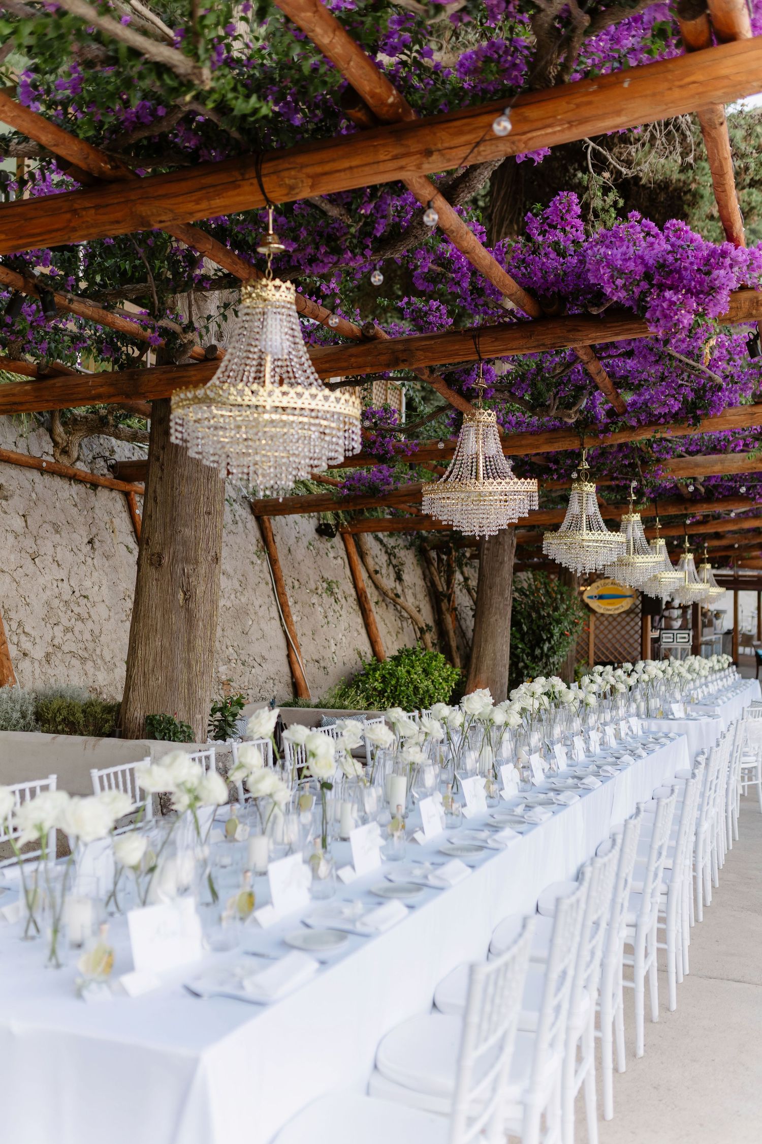Elegant outdoor wedding reception with crystal chandeliers, long white tables, and purple bougainvillea draped over a wooden pergola.