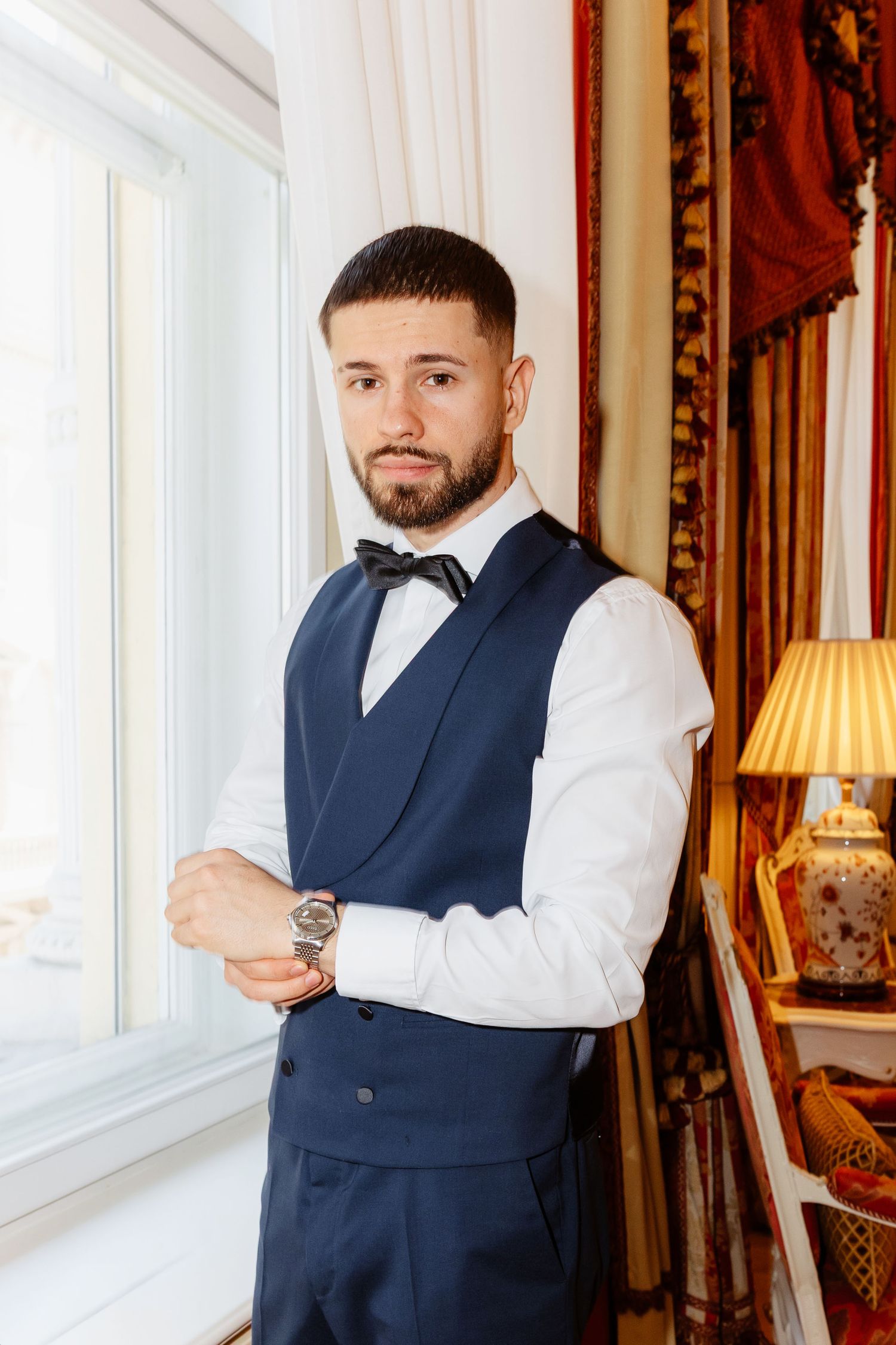 Elegant groom in navy blue vest and bow tie adjusting his watch before wedding ceremony.