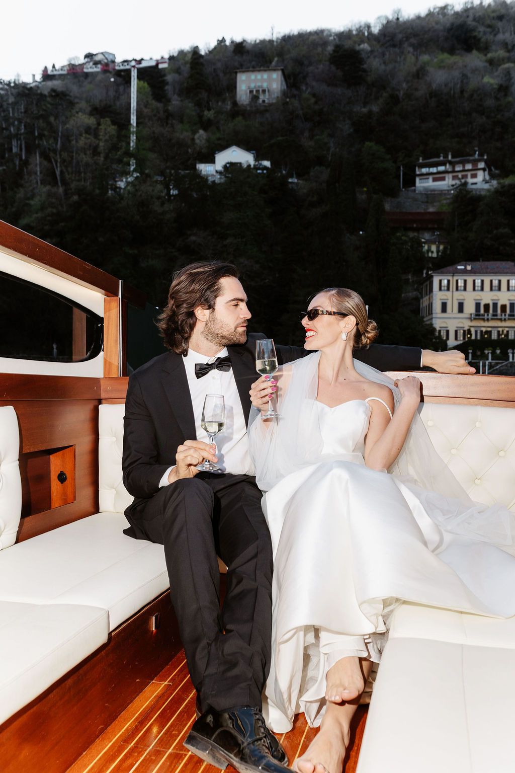 Bride and groom in wedding attire sipping champagne on a luxury yacht with scenic hillside backdrop.